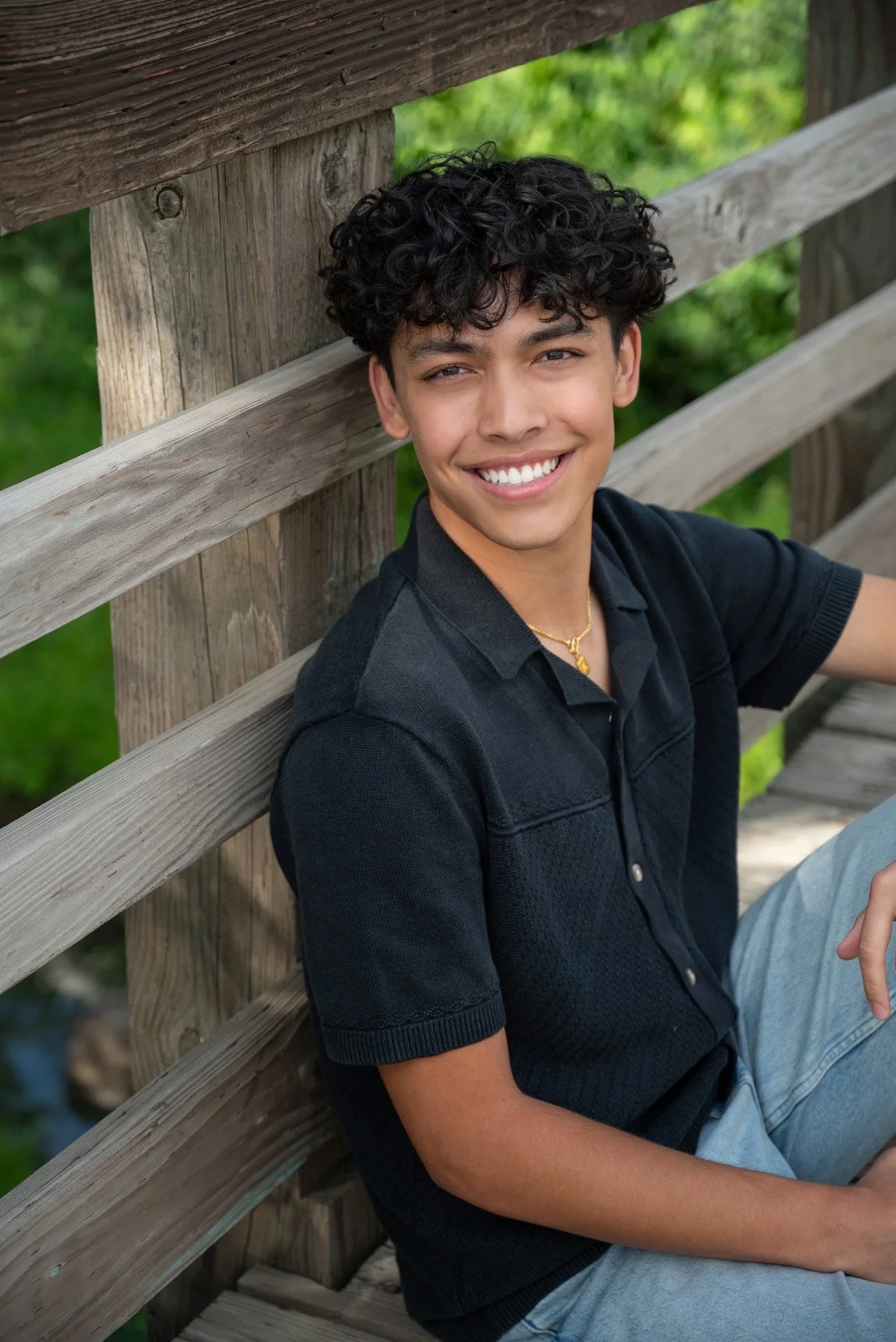 A young man with curly black hair smiling and sitting on a wooden park bench with a green leafy background.