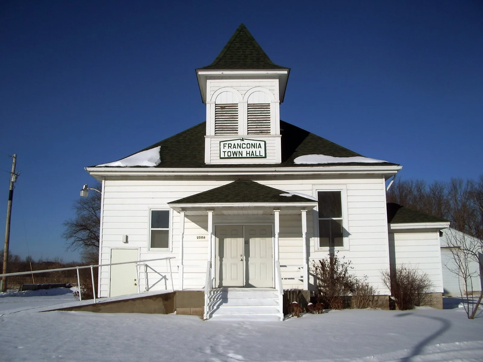 White town hall building with a peaked roof, a bell tower, and a sign that reads 'Franconia Town Hall'. Snow on the roof and ground, blue sky in the background.