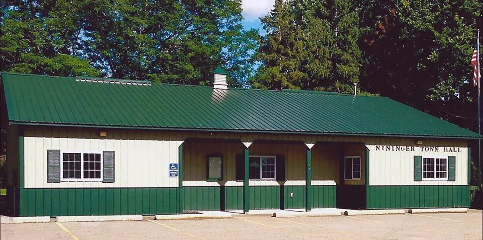 A small town hall building with a green metal roof and green and white outside walls, three windows with black shutters, a handicapped parking sign, and a parking lot in front. The building has a sign that reads 'NININGER TOWN HALL' and an American f