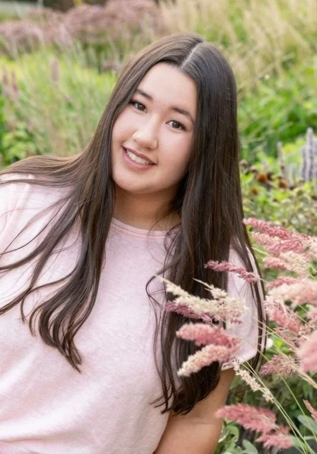 A young woman with long brown hair smiling outdoors among pink and green plants.