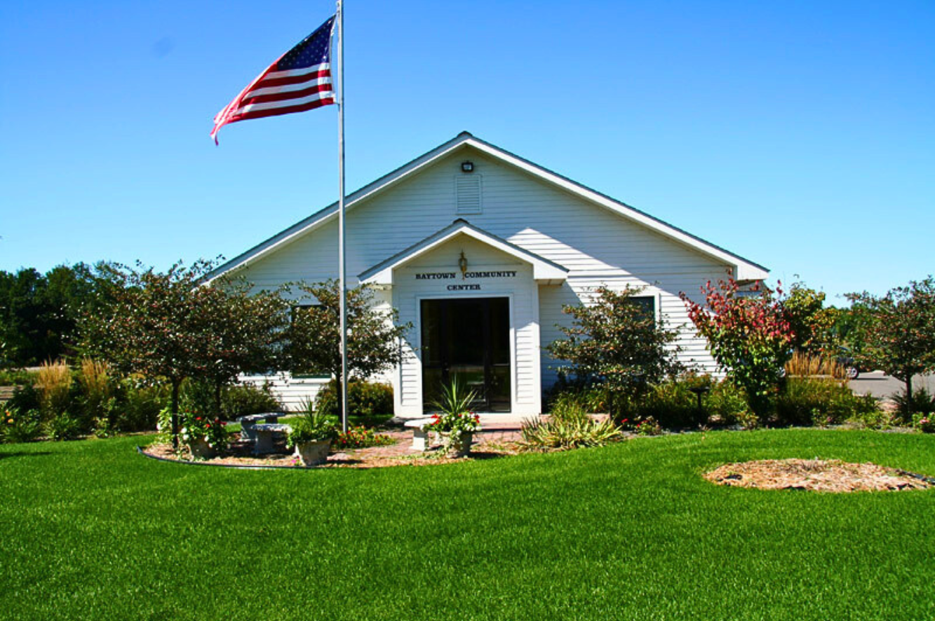 Front view of a white community center building with a sign that reads 'Kaytown Community Center' under a clear blue sky, surrounded by landscaped garden and American flag on a flagpole.