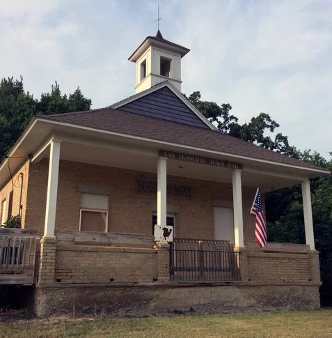 A small church building with a brick foundation, white columns, a blue roof, and a small steeple on top. An American flag hangs on the right side of the porch.