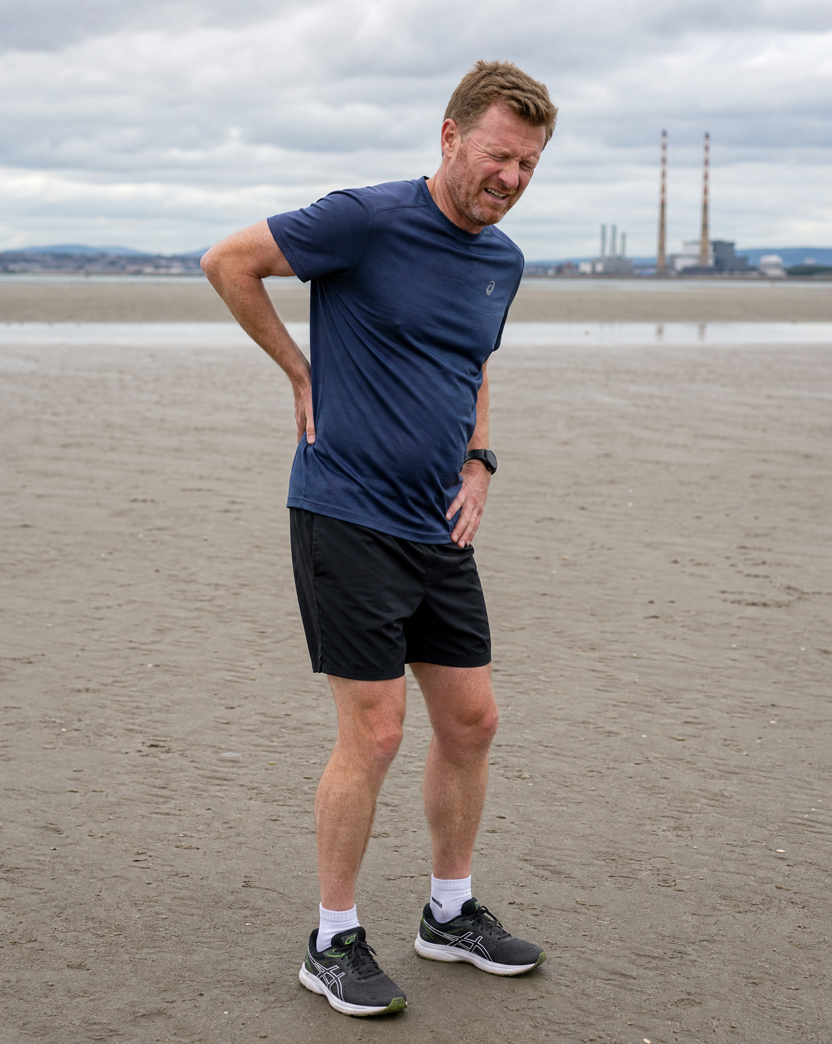 A man in athletic clothing standing on a beach with his hand on his back and a pained expression, with industrial buildings and cloudy sky in the background.