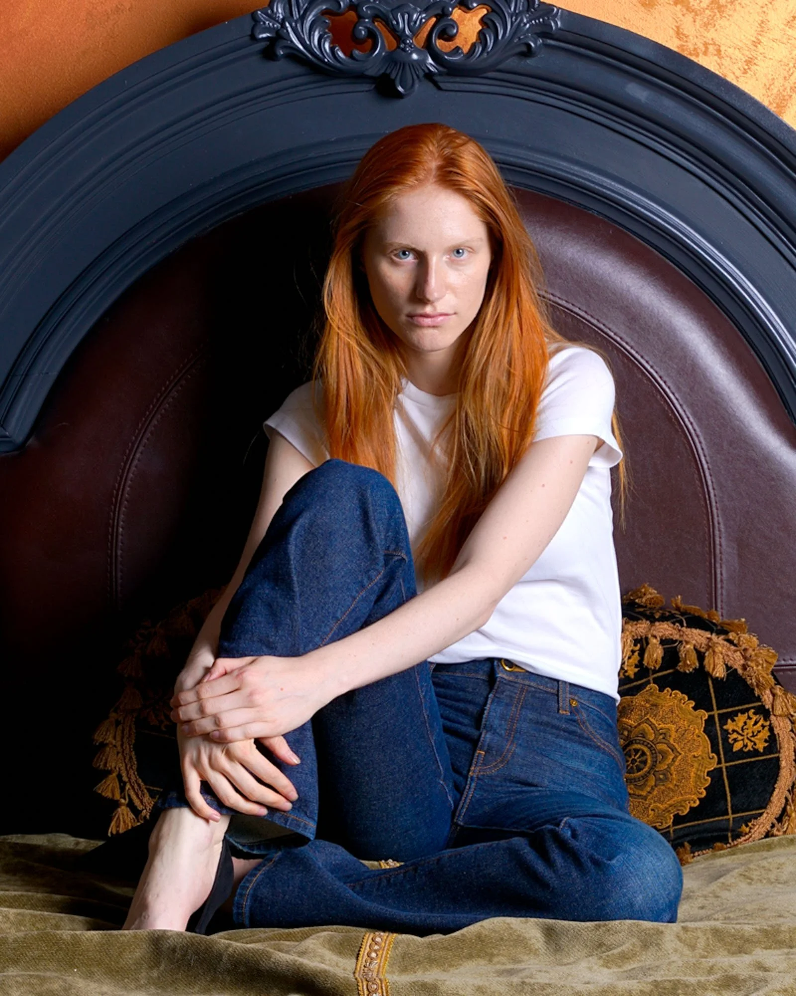 A young woman with long red hair, light skin, and blue eyes, sitting cross-legged on a tan-colored bed. She is wearing a white t-shirt and blue jeans. She is looking directly at the camera with a neutral expression. Behind her is a large, dark, ornat