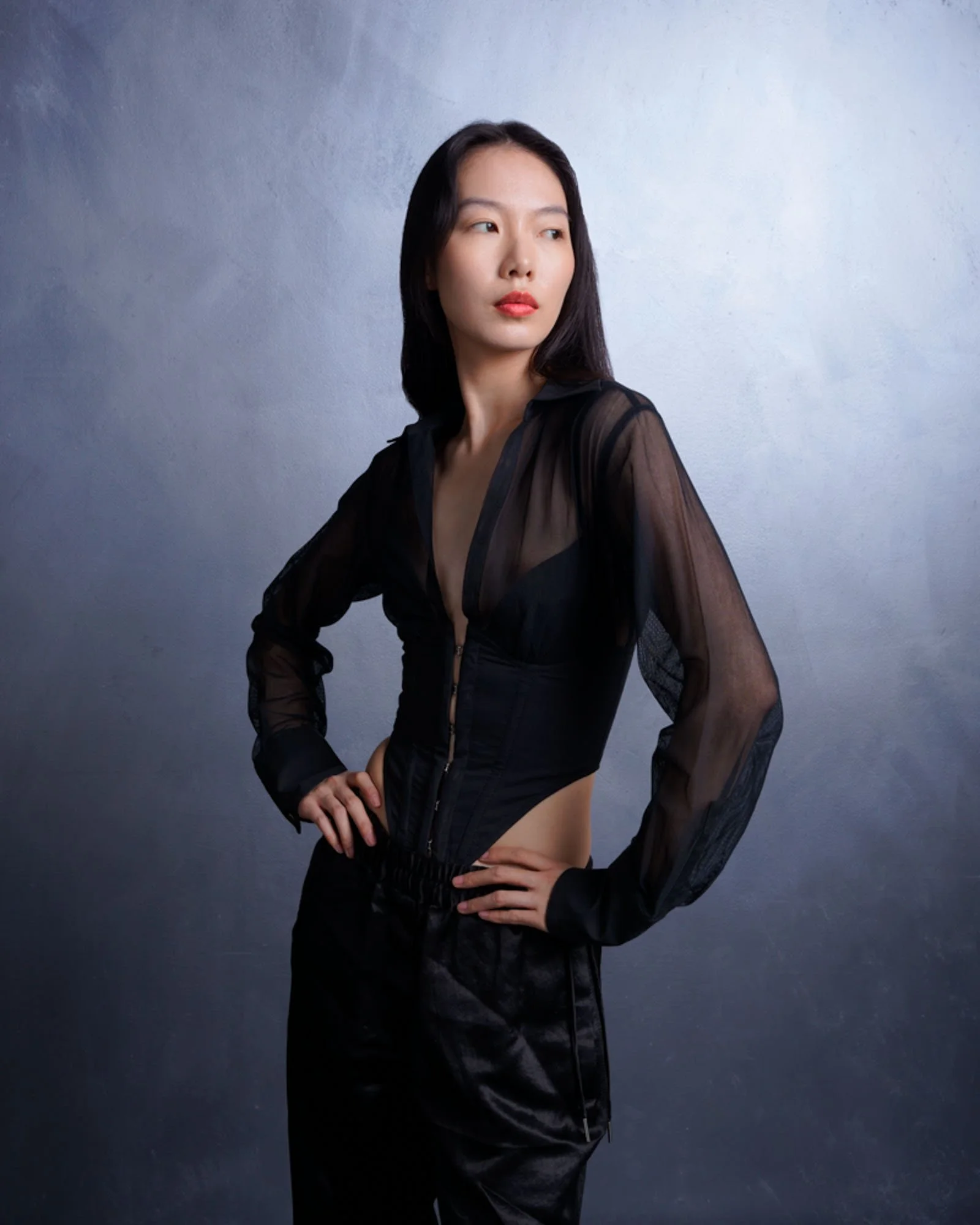 A woman with long dark hair wearing a black sheer jacket and black pants, posing against a light blue wall.