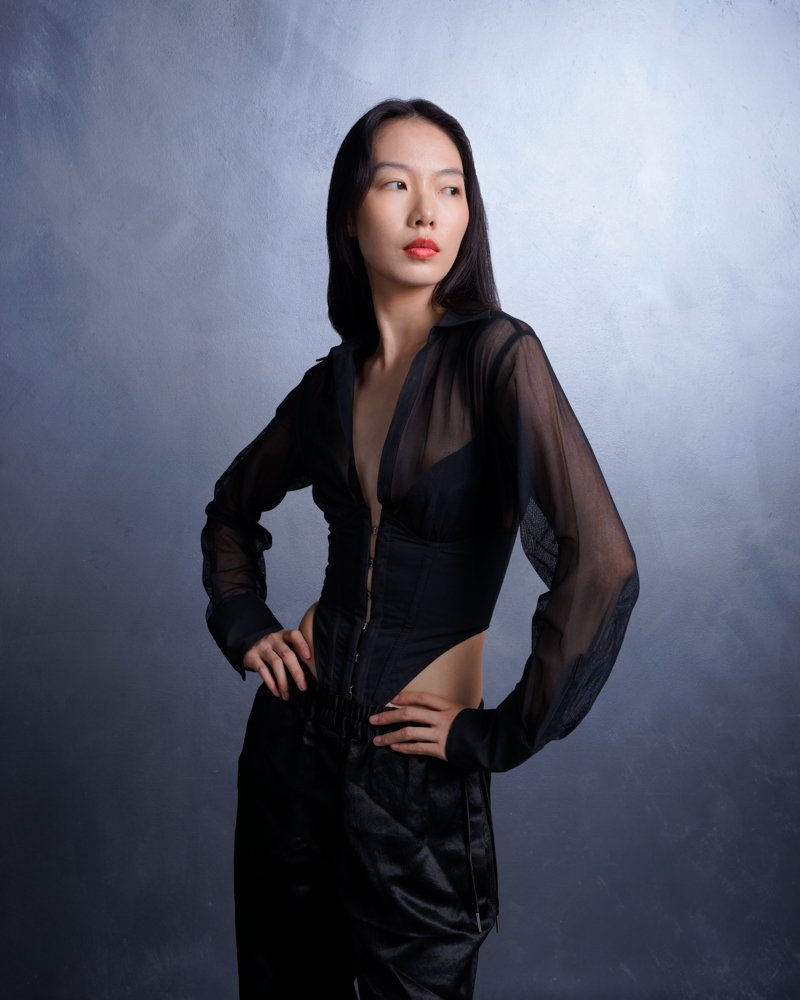 A woman with long black hair wearing a sheer black jacket and black pants, standing against a neutral background.