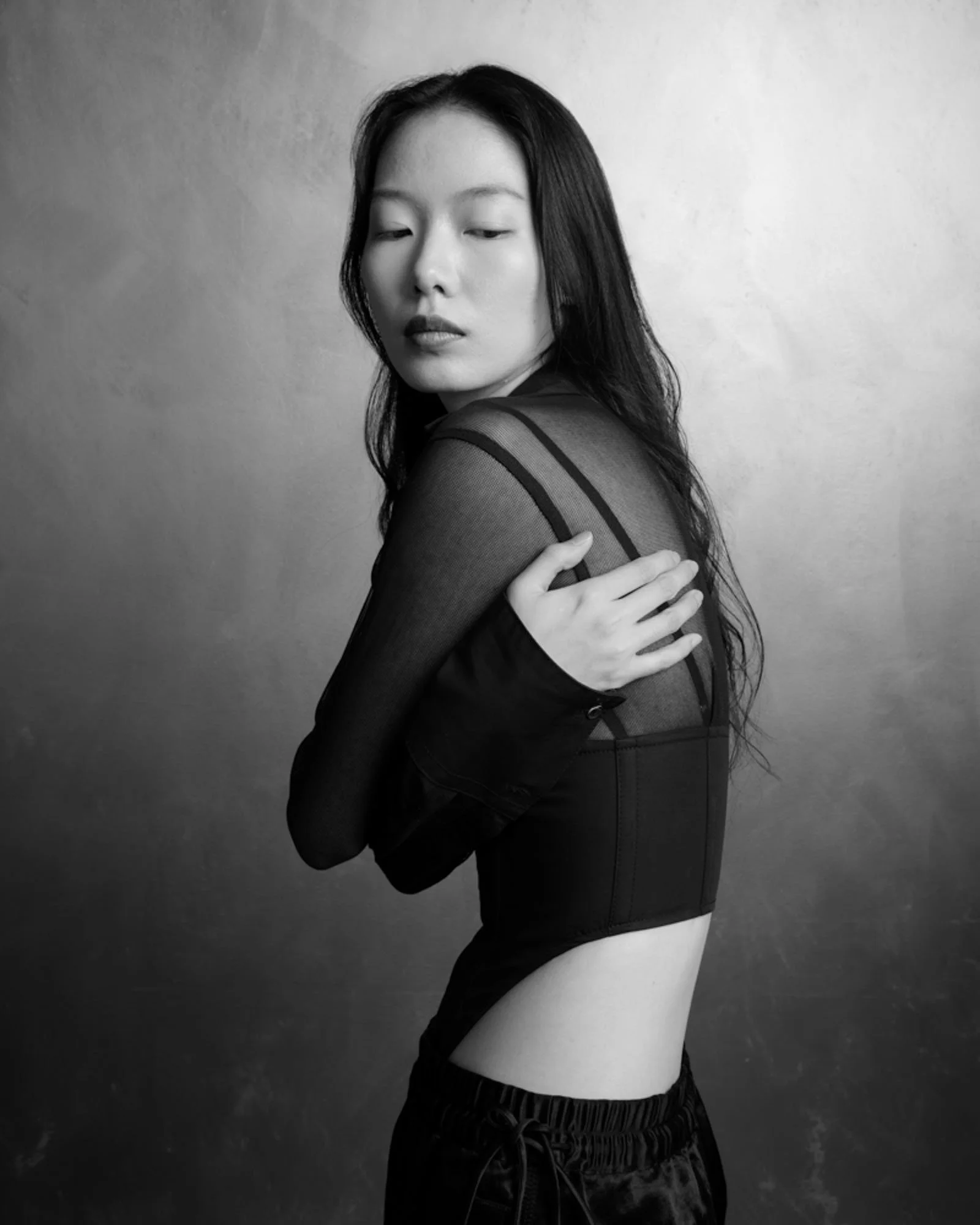 Black and white photo of a woman with dark, wavy hair wearing a sheer, long-sleeved top and black shorts, standing against a plain background.