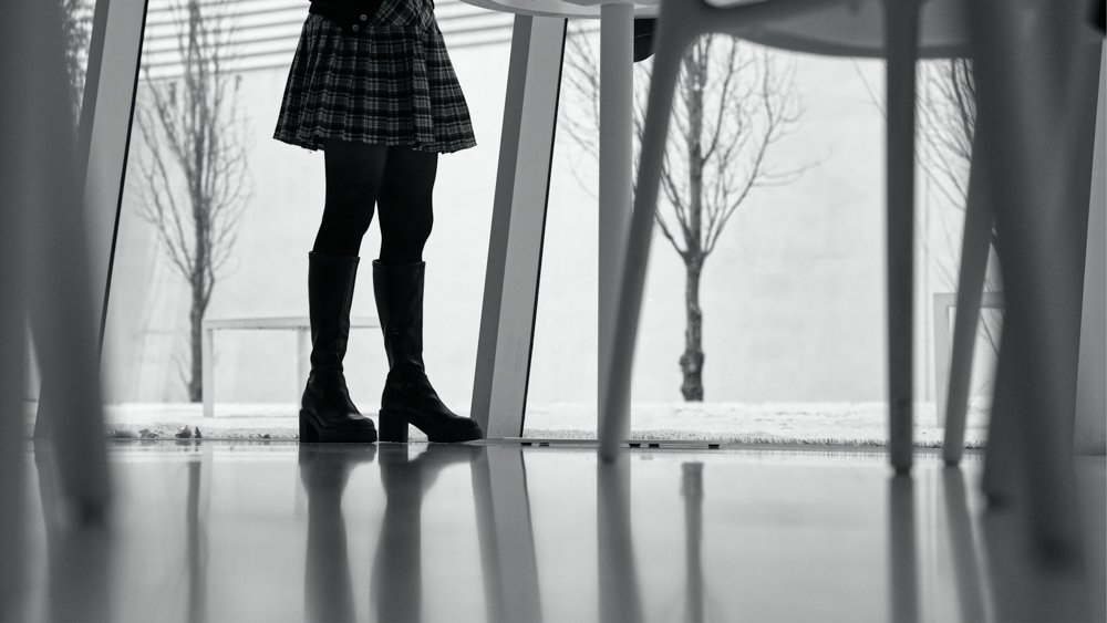 Person standing indoors, shot from below, wearing a plaid skirt, black tights, and black boots, with windows and leafless trees visible outside.