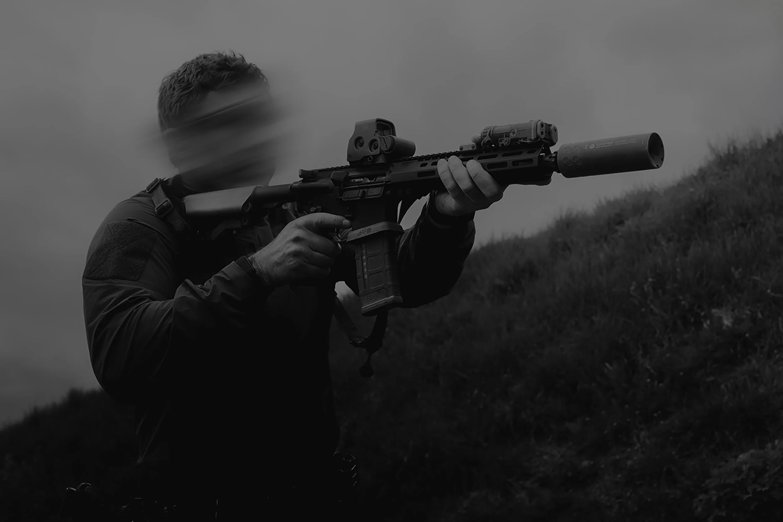A person aiming a rifle outdoors, wearing tactical gear and protective eyewear, with a smoke-filled sky in the background.
