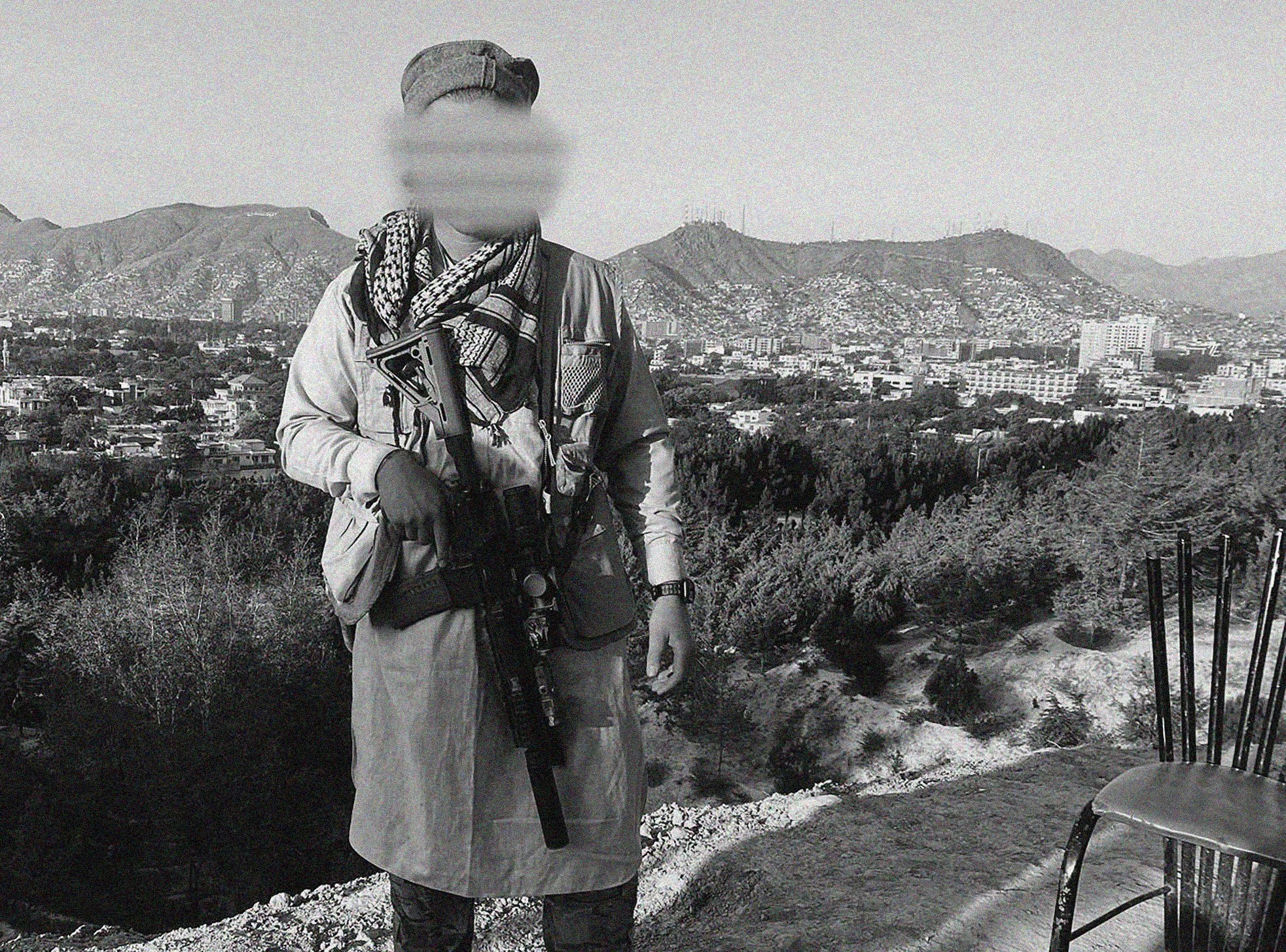 A person wearing a headscarf, jacket, and sunglasses standing outdoors on a hill with a cityscape and mountains in the background, holding a weapon.