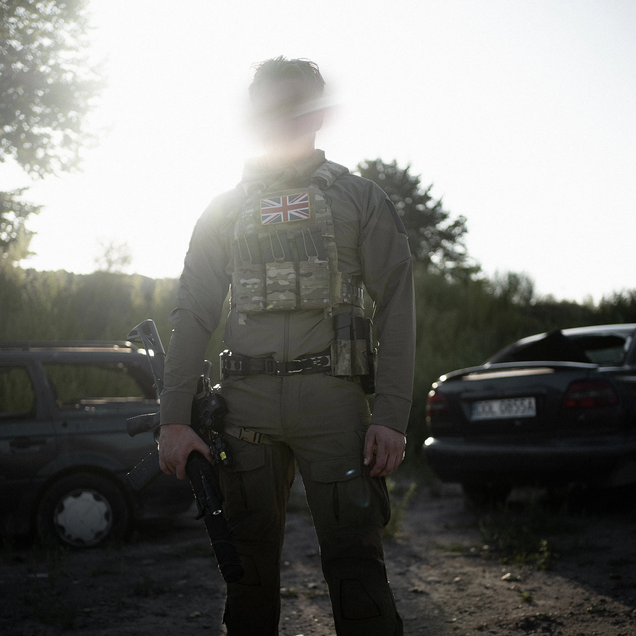 A person dressed in tactical gear with a British flag patch on their chest, standing outdoors during daylight, with vehicles and trees in the background.