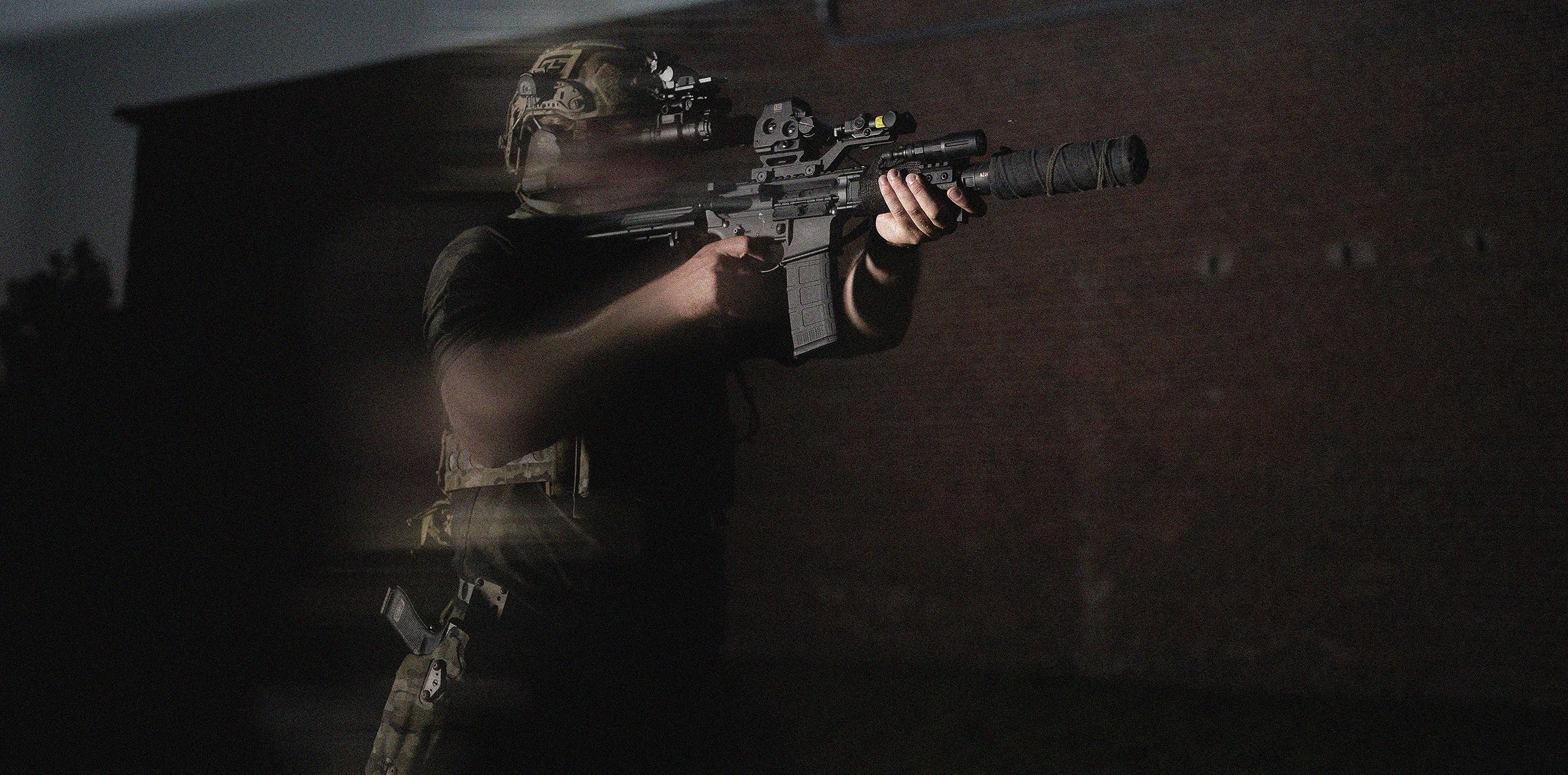 A soldier wearing camouflage gear and a helmet, aiming a scoped rifle with a suppressor attached, against a dark background.