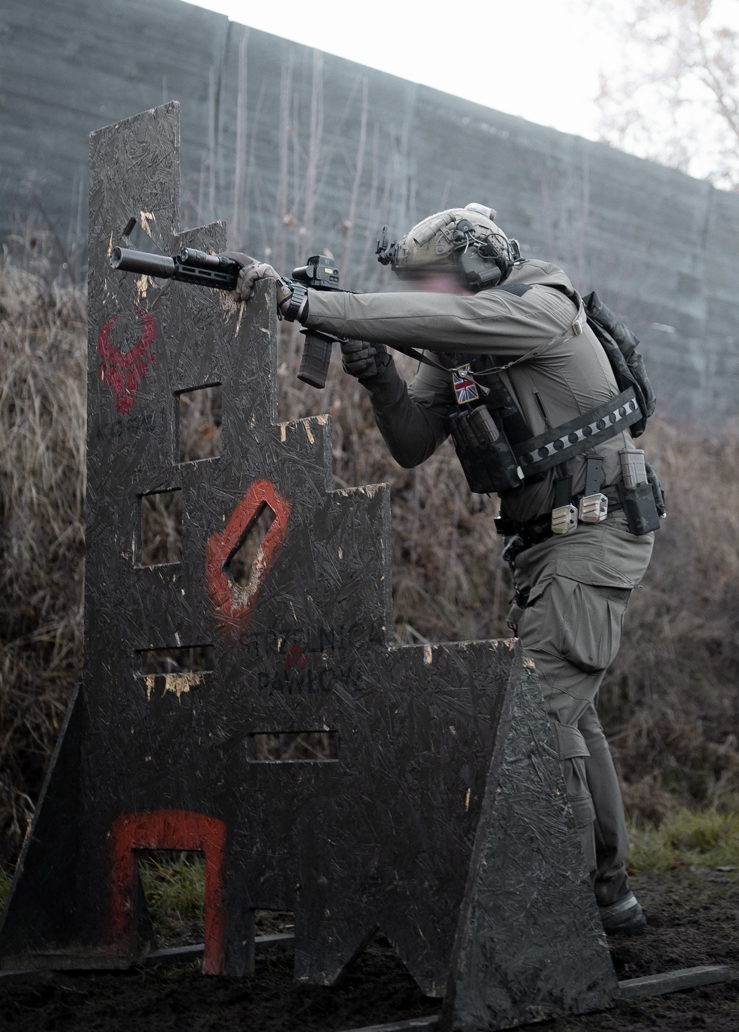 A police officer wearing tactical gear aiming a rifle over a wooden barricade with red markings, outdoors with a wall and trees in the background.