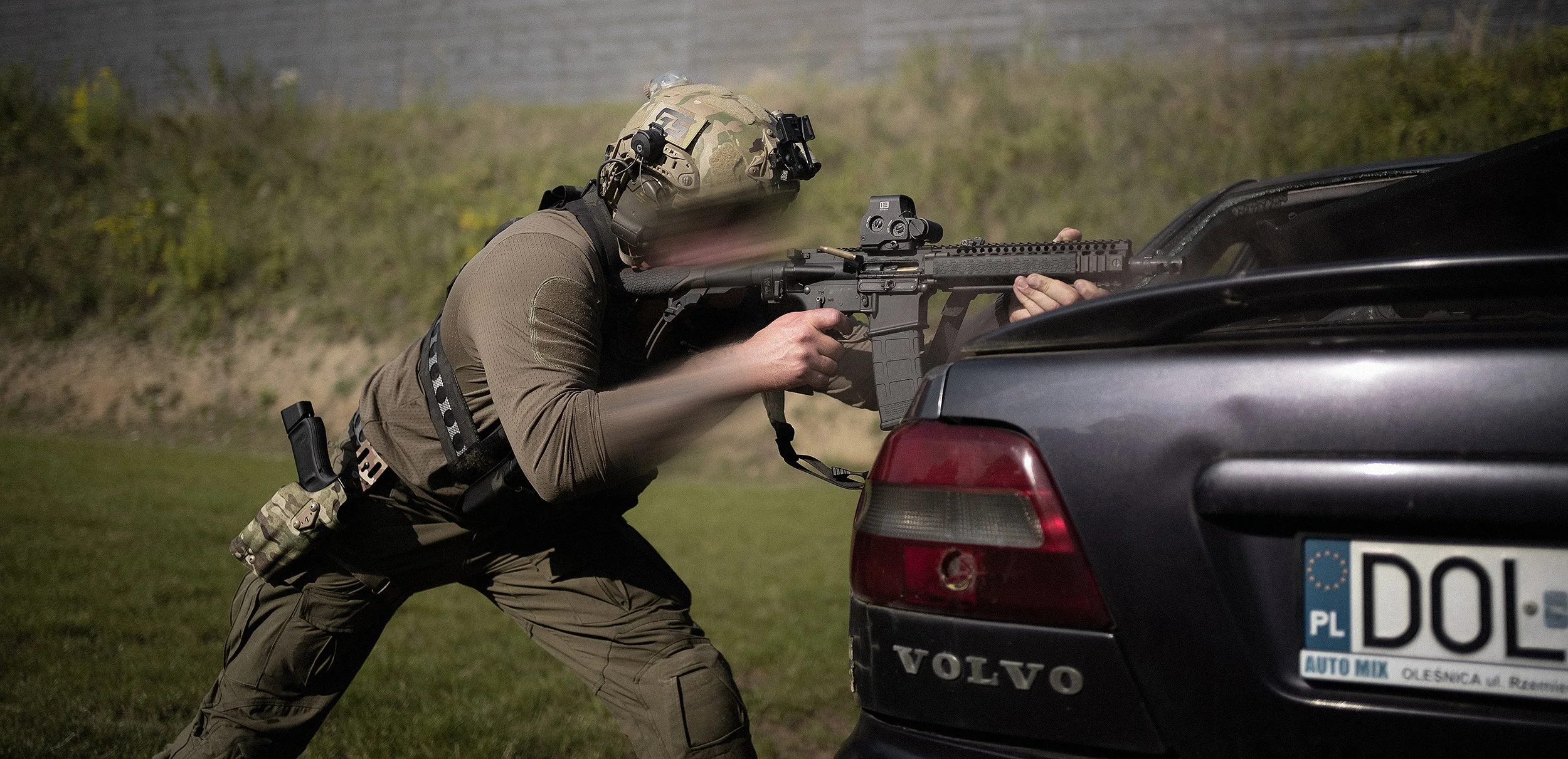 A soldier in tactical gear aiming a rifle from the back of a police car, with a European license plate, in an outdoor setting with grass and bushes in the background.