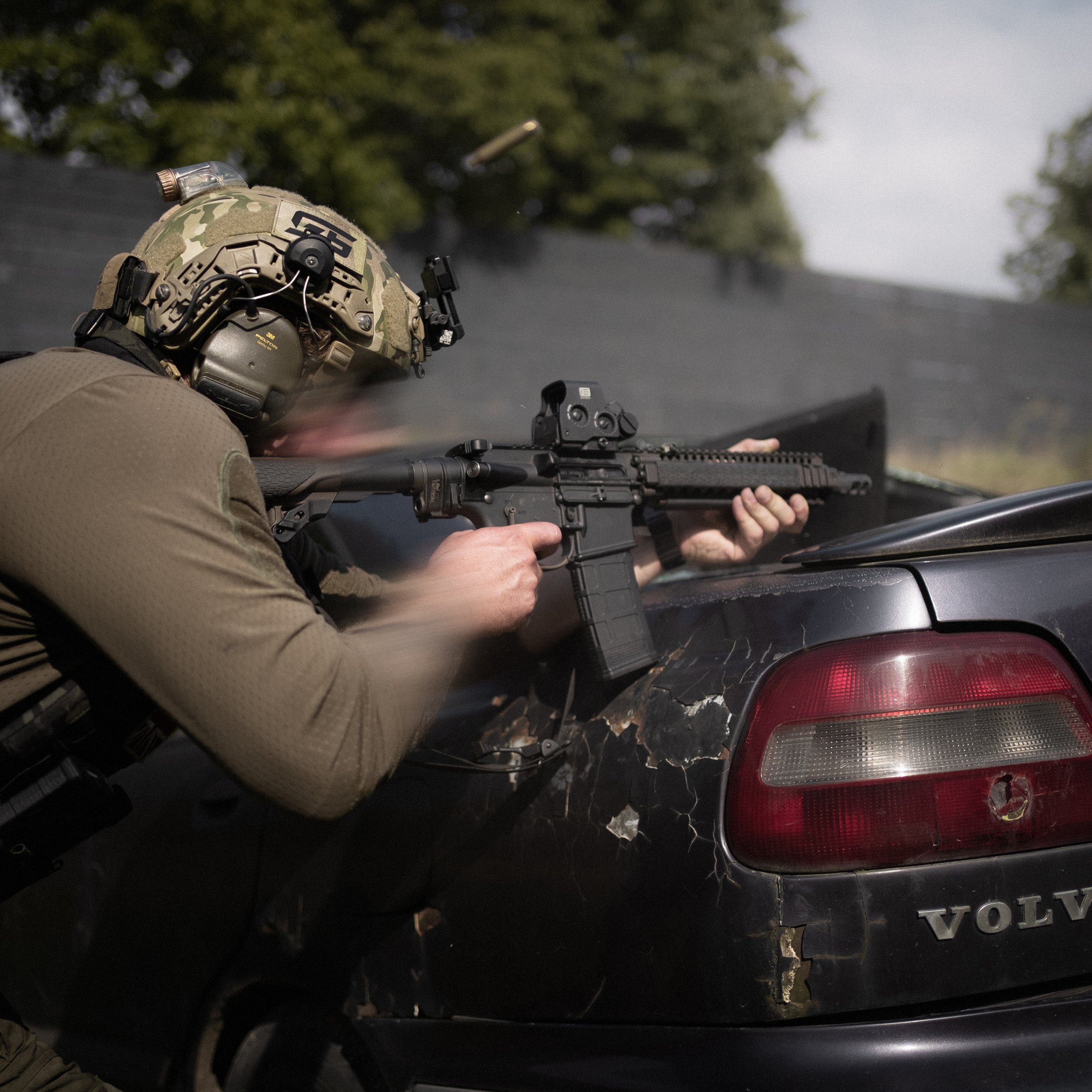Person in military gear aiming an assault rifle from the back of a vehicle with a partially visible Volvo logo and rear tail lights.