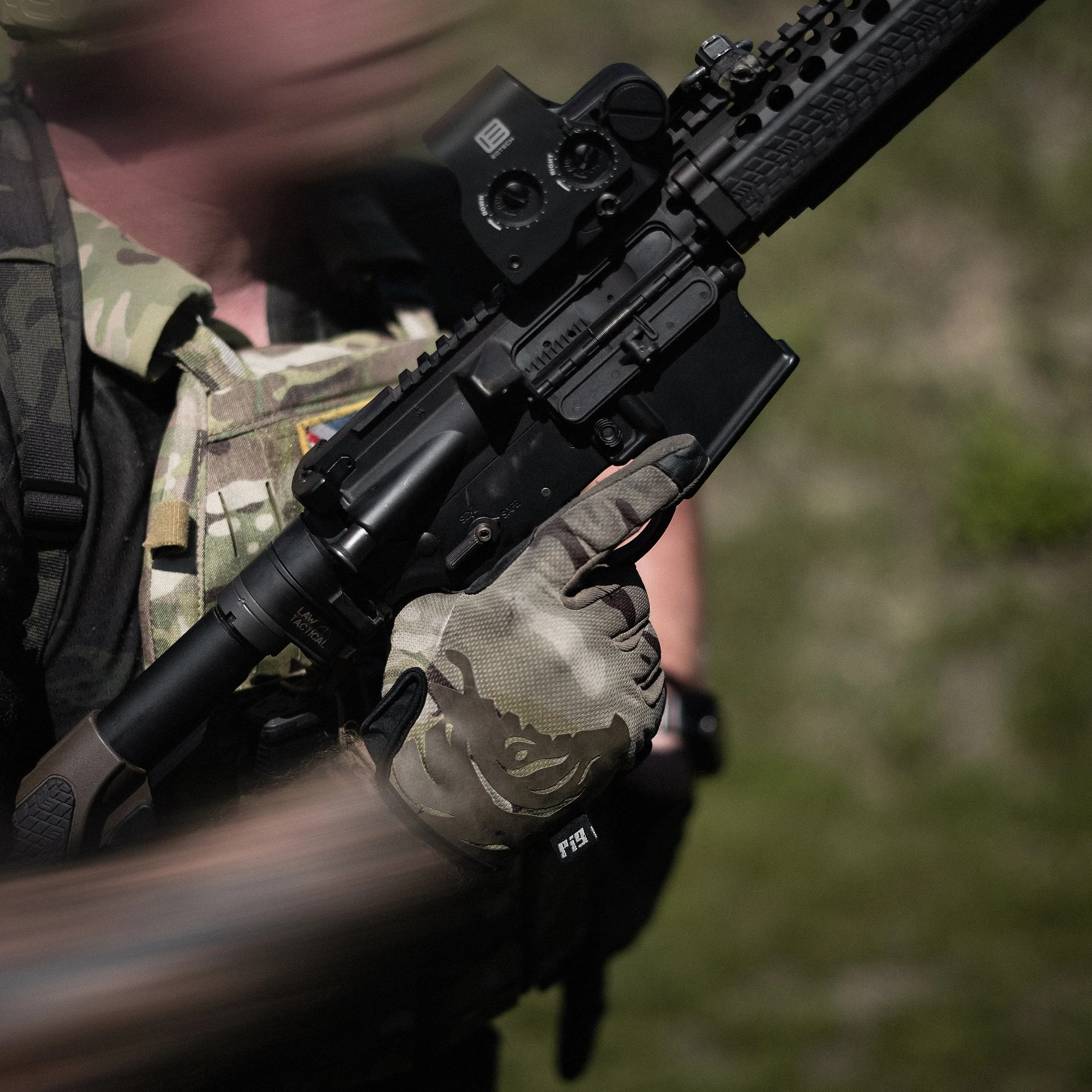 Close-up of a person holding a black firearm with a tactical rail, wearing camouflage clothing.