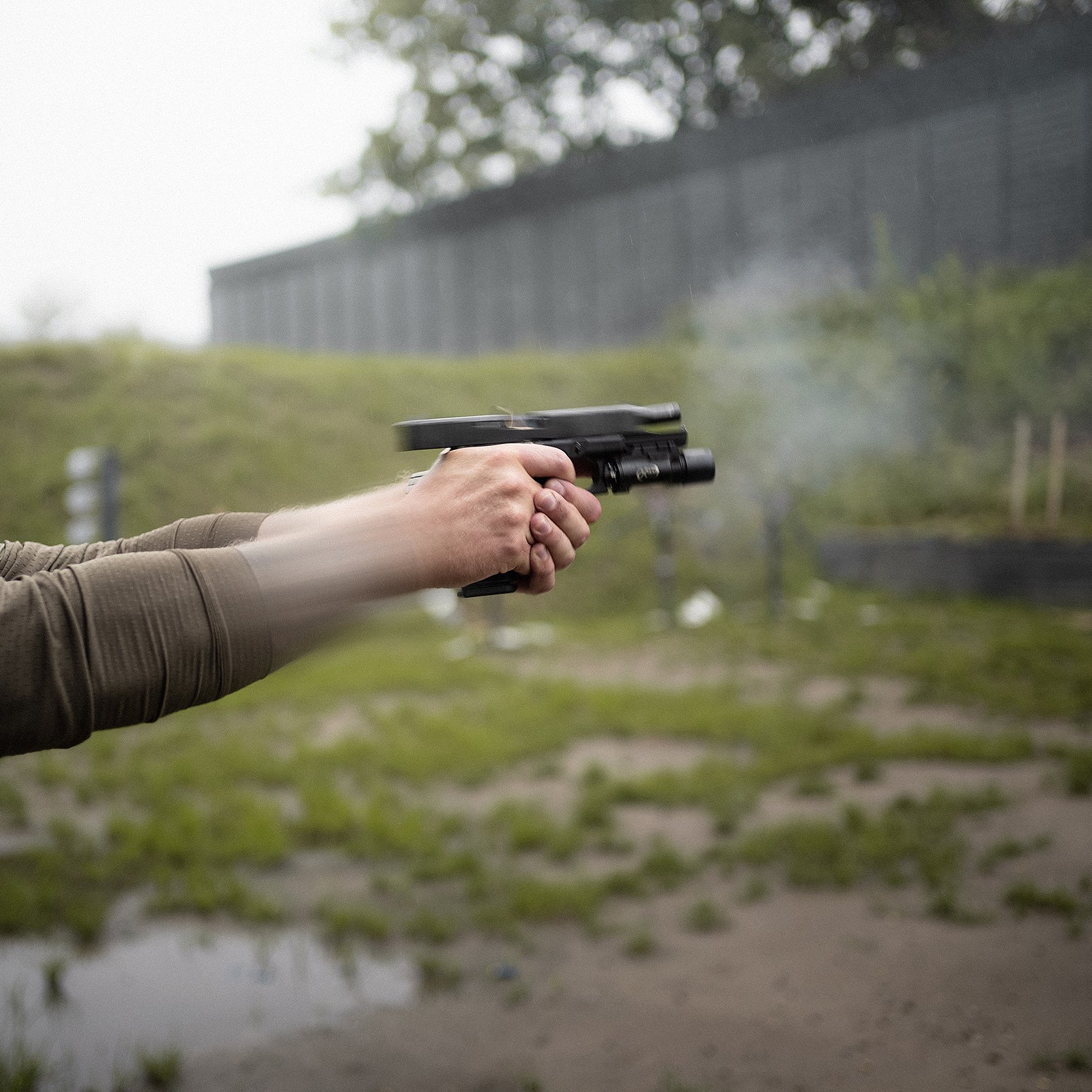Person shooting a handgun outdoors with a grassy field and cloudy sky in the background.