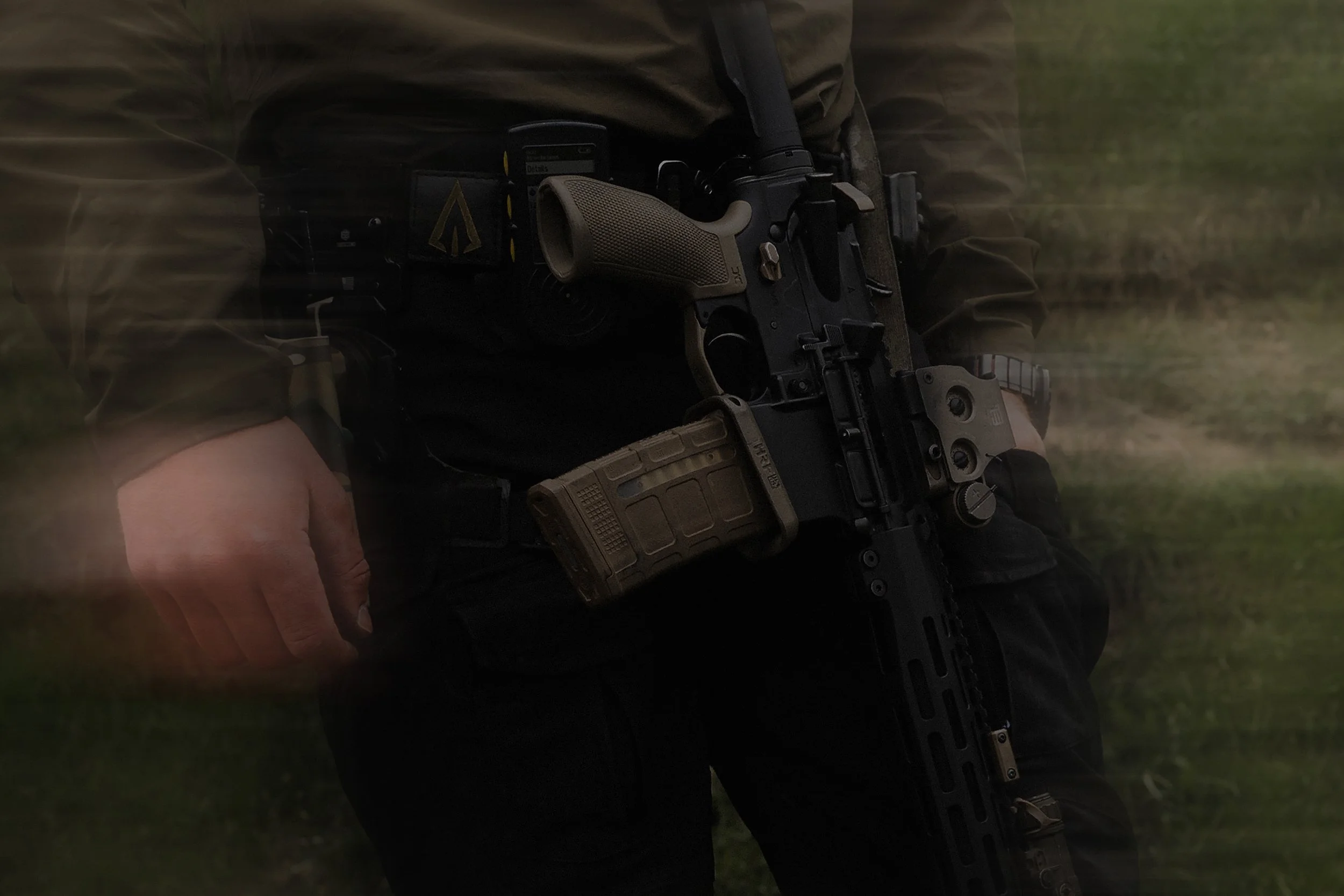 A person holding a tactical firearm with a tan grip, attached to tactical gear, in an outdoor setting.
