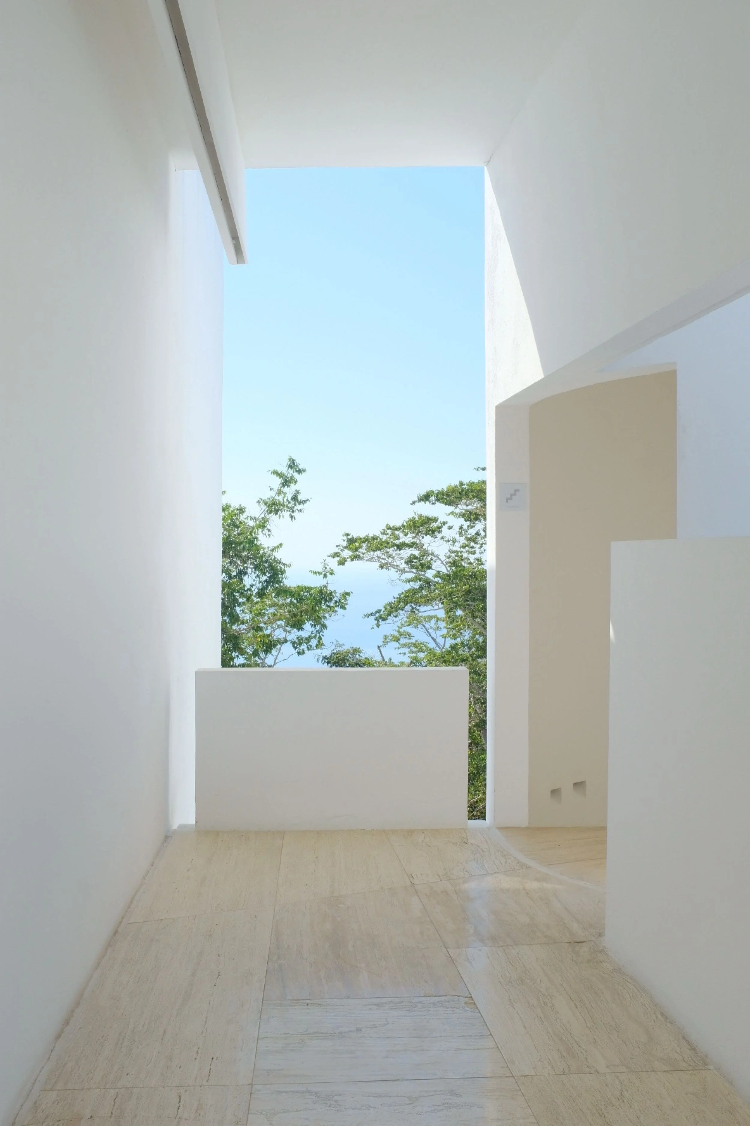 Pasillo minimalista de muros blancos de Miguel Ángel Aragonés abriéndose hacia la vegetación tropical.
