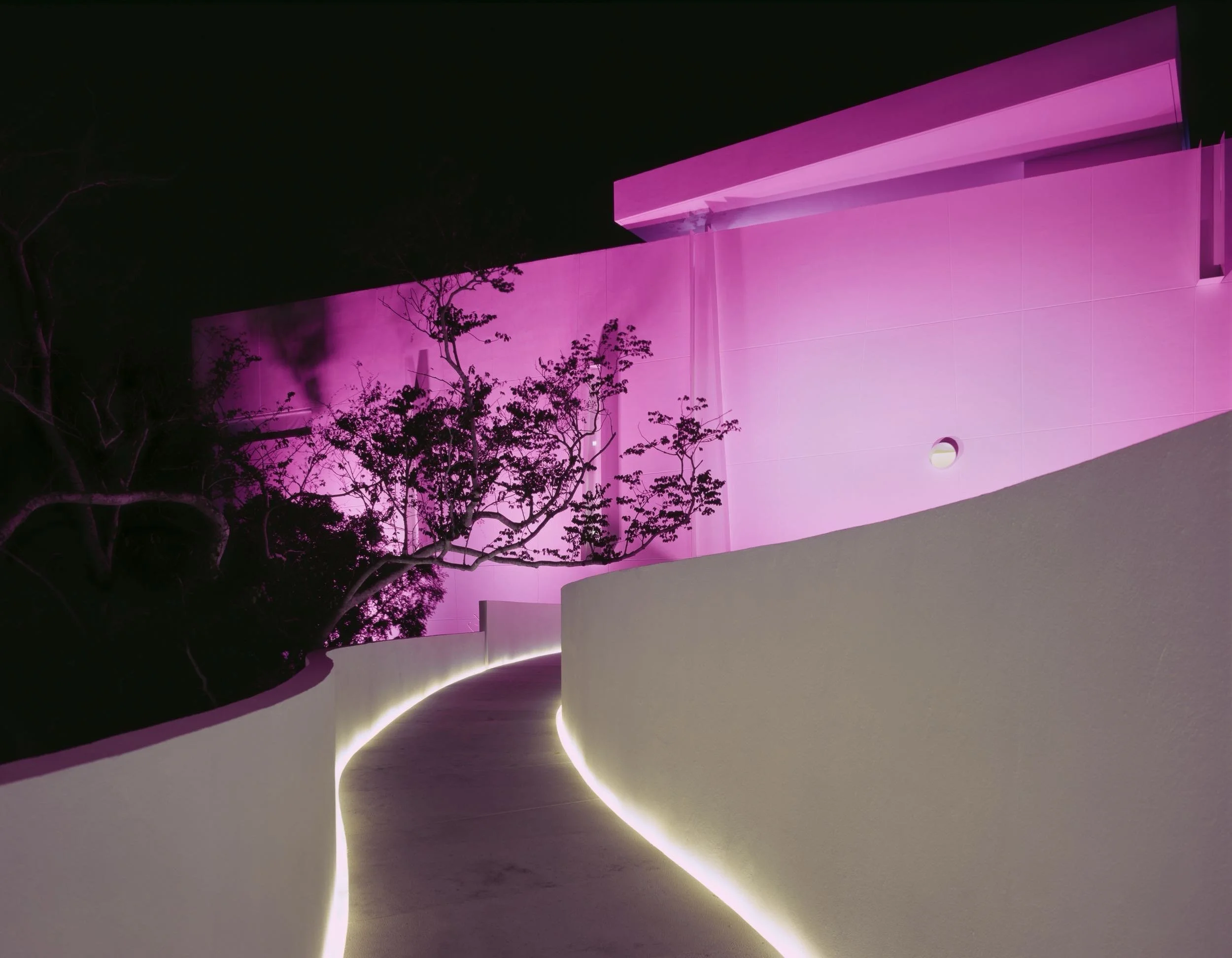 Iluminación artística nocturna y diseño cromático en las instalaciones de lujo de Hotel Encanto Acapulco. Fotografía de Joe Fletcher. Pasillo exterior haci estructura en color magenta.