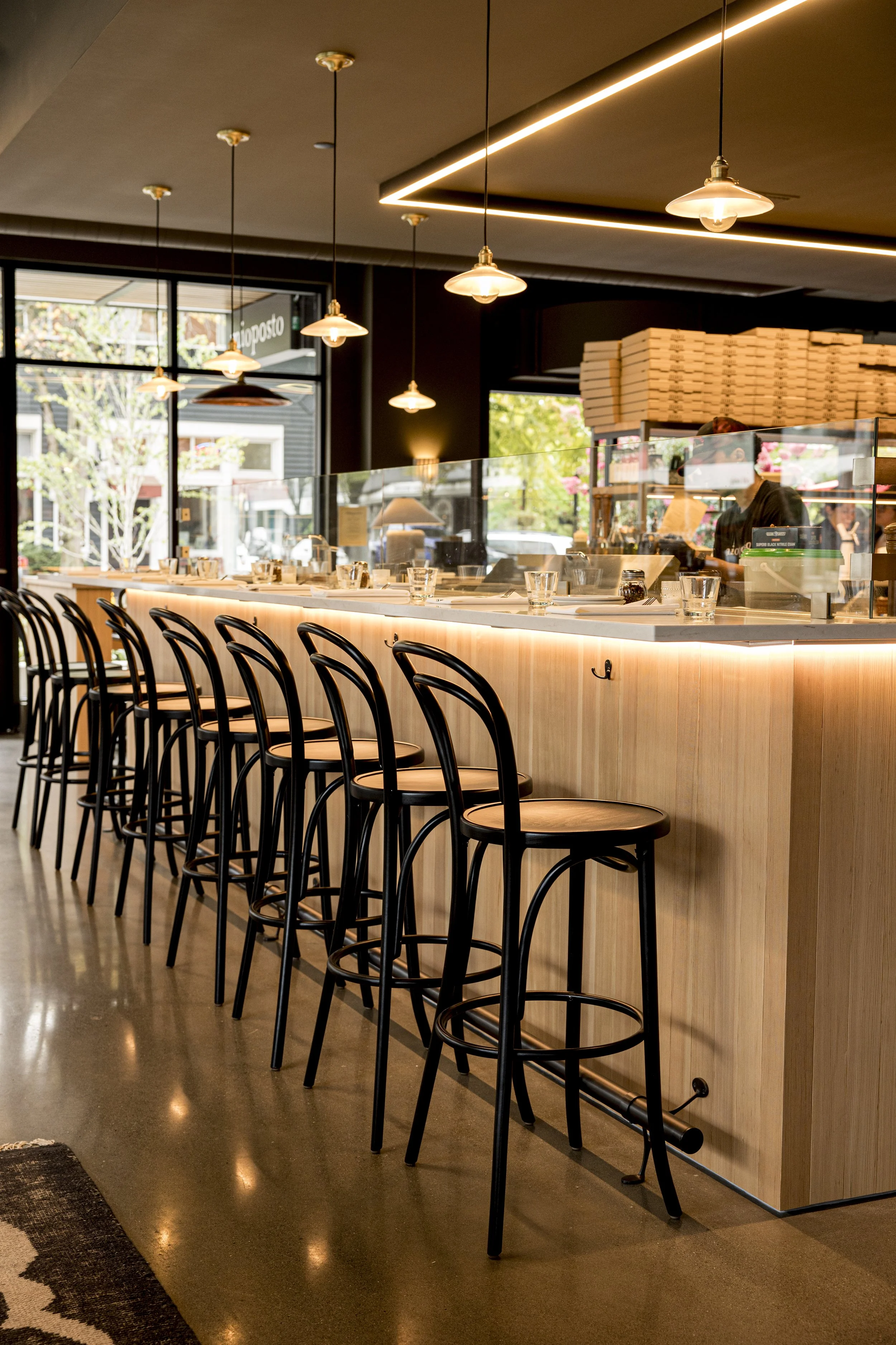 Interior of a modern restaurant with a wooden counter, black chairs, pendant lights, large windows showing trees and buildings outside, and a stack of pizza boxes.