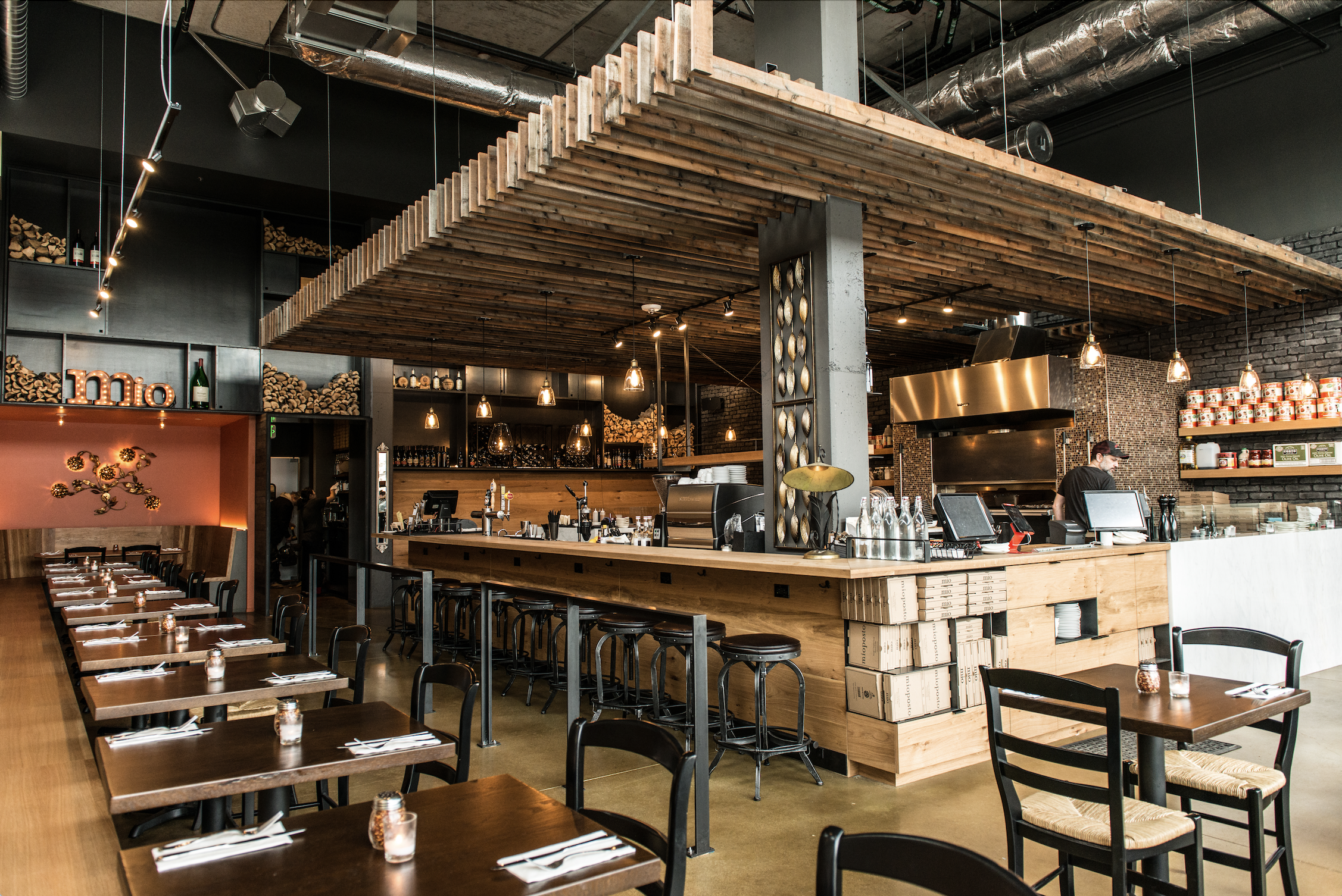 Inside a modern restaurant with wooden tables, black chairs, and a bar area with hanging lamps, wood accents, and a brick wall.