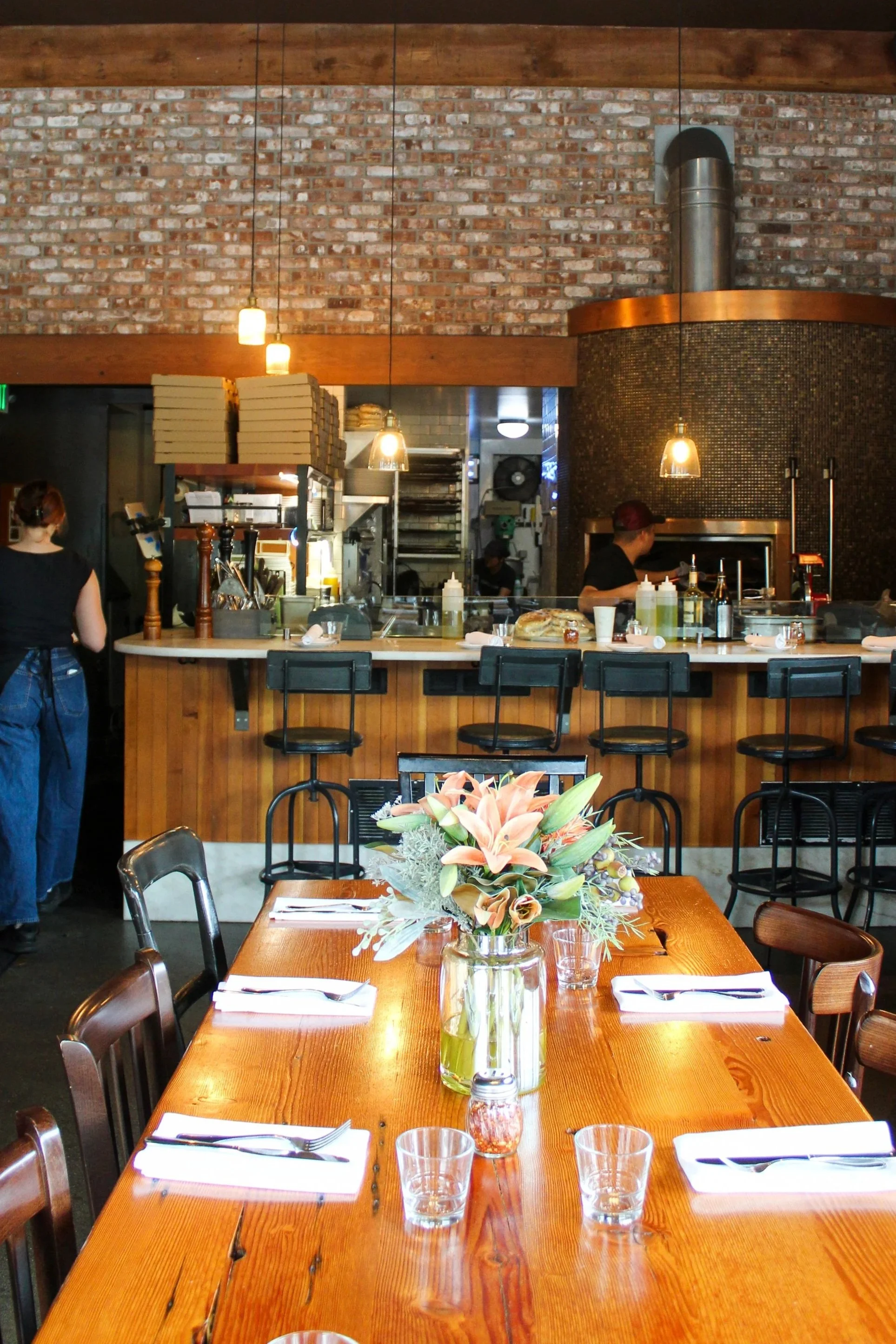 Interior of a restaurant with a wooden dining table, flower centerpiece, and set tables. In the background, there is a bar counter, kitchen area, and brick wall with hanging lights.
