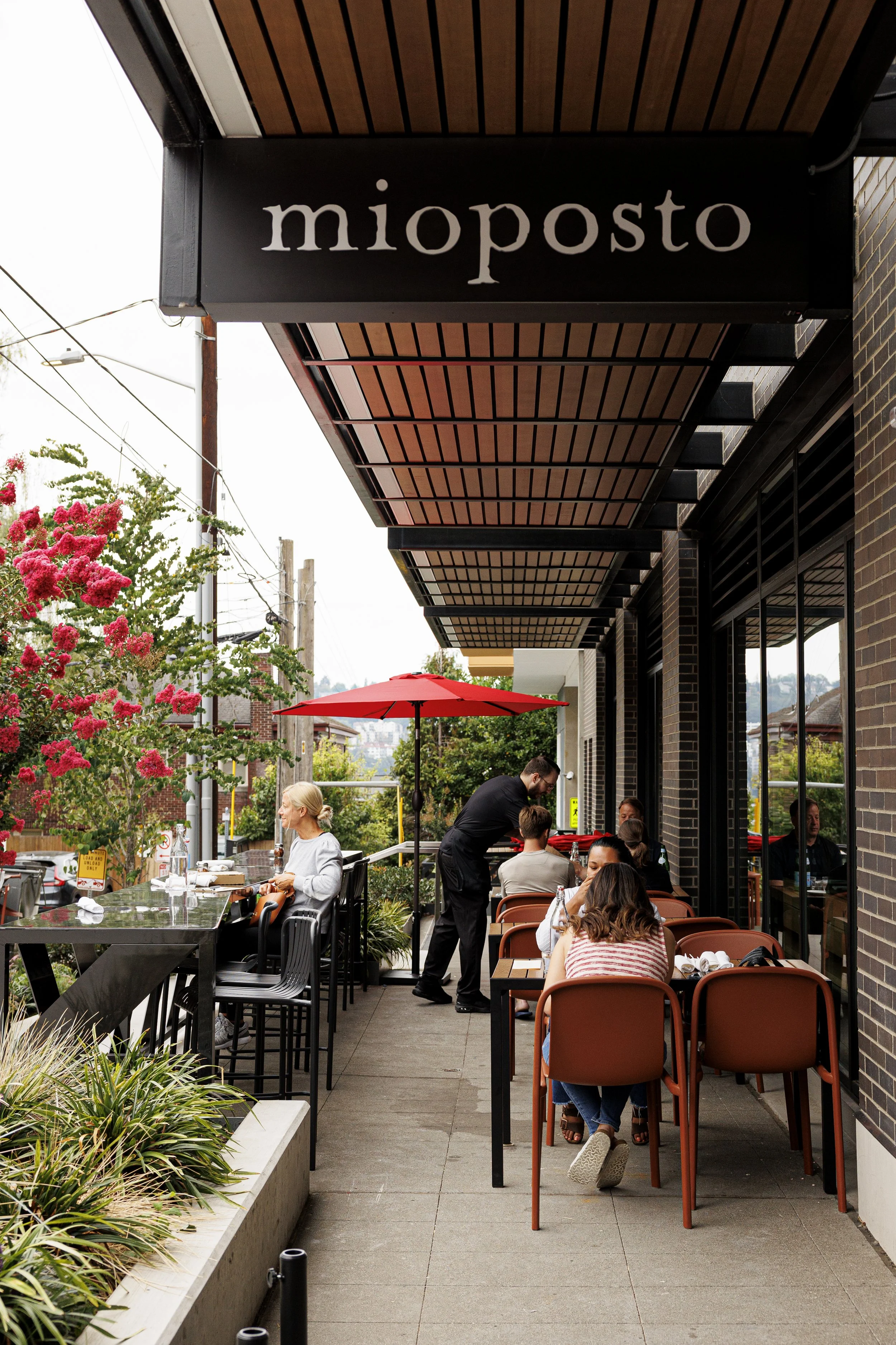 Outdoor dining area of a restaurant with several tables and chairs, some tables with customers. A black sign with white text reads 'mioposto' above the entrance. People are sitting and conversing, and a waiter is attending to a table. There are flowe