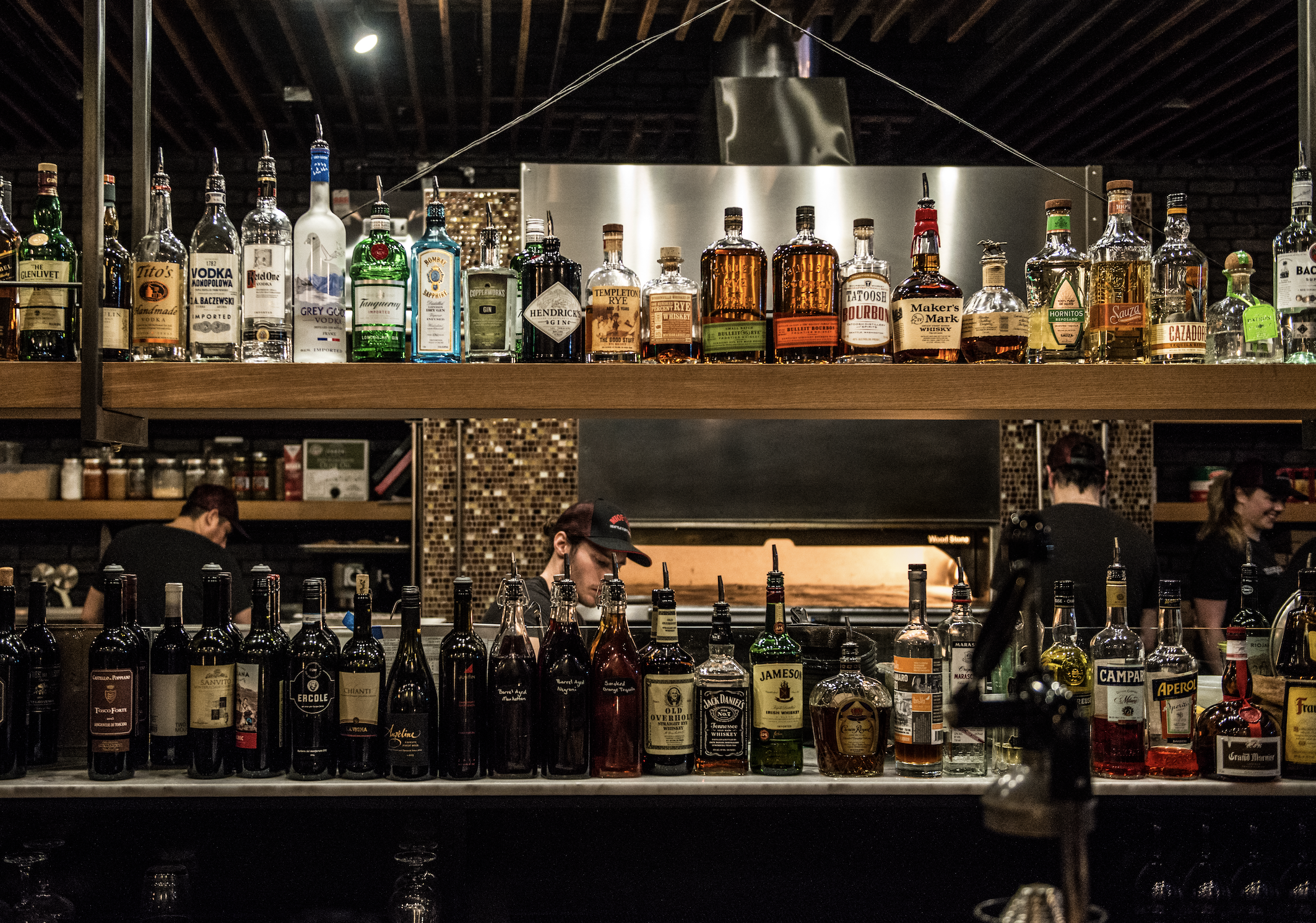 A bar shelf displaying various bottles of liquor, including whiskey, vodka, and gin, in a dimly lit restaurant or bar setting. People are working behind the bar, and a person in the foreground is partially visible.