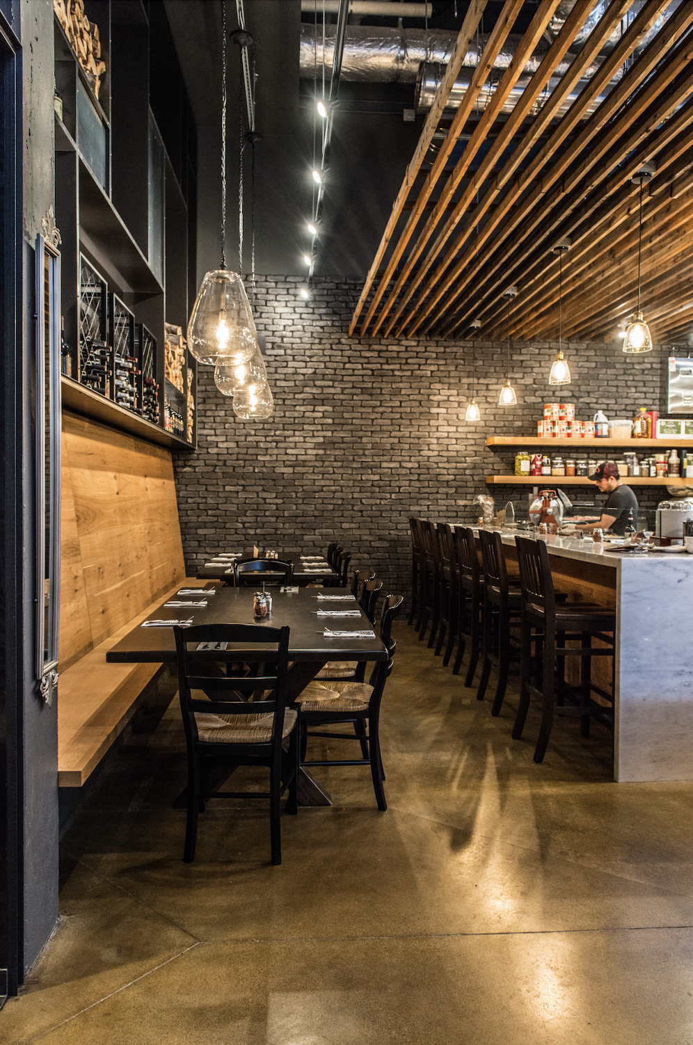 Inside a modern restaurant featuring a brick wall, wooden ceiling slats, black tables with chairs, and hanging glass pendant lights. A bar area with high chairs and shelves with food products and seasonings is visible, along with a person preparing f