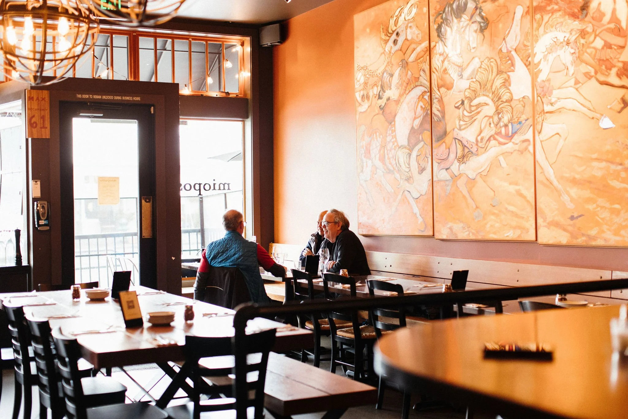 Interior of a restaurant with tables, chairs, and large wall art featuring horses, riders, and Native American motifs. Two men are seated at a table near a large window, engaged in conversation.