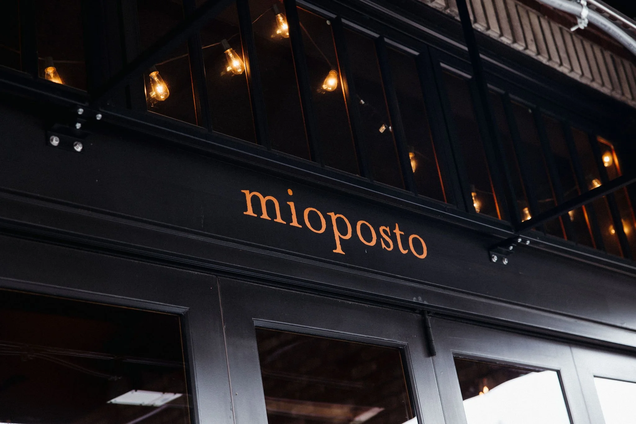 Close-up of a black storefront with the name 'miopost' in orange lettering, illuminated through large glass windows with visible interior light bulbs, at night.
