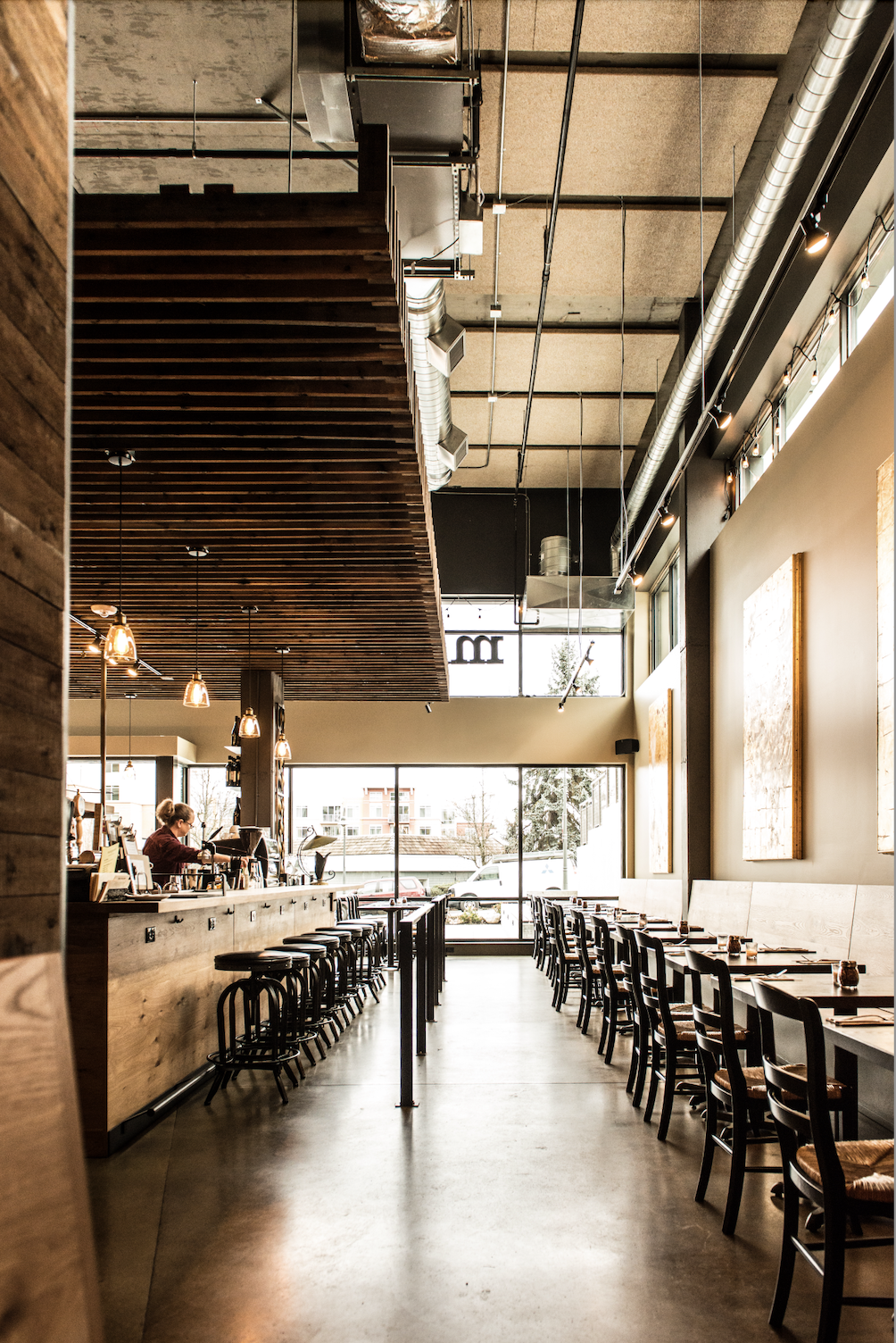 Interior of a modern restaurant with a bar on the left, wooden ceiling accents, and large windows at the back, with tables and chairs on the right.