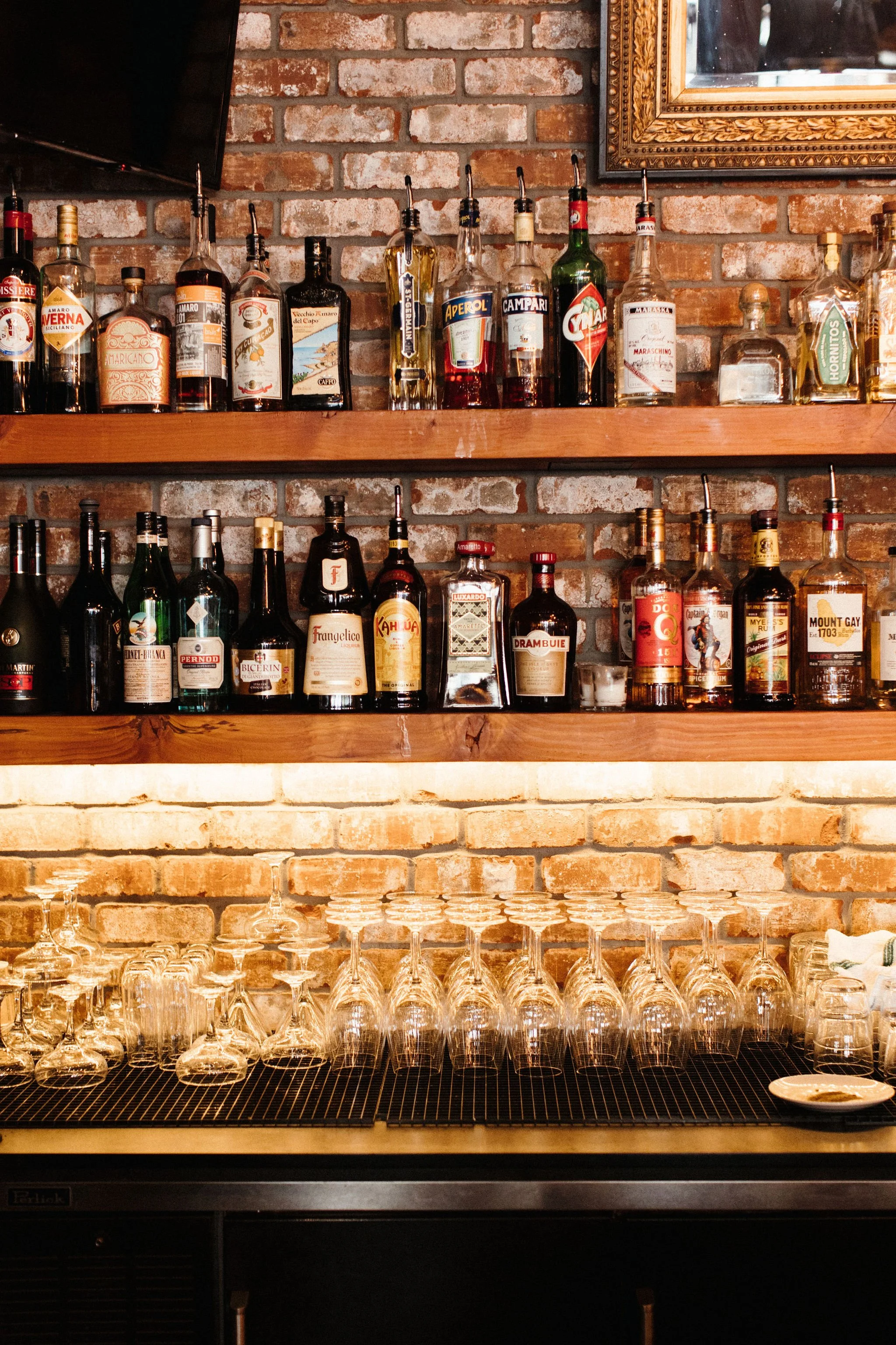 Multiple bottles of alcohol on a wooden shelf against a brick wall, with glassware and a mirror reflecting part of the bar setup.