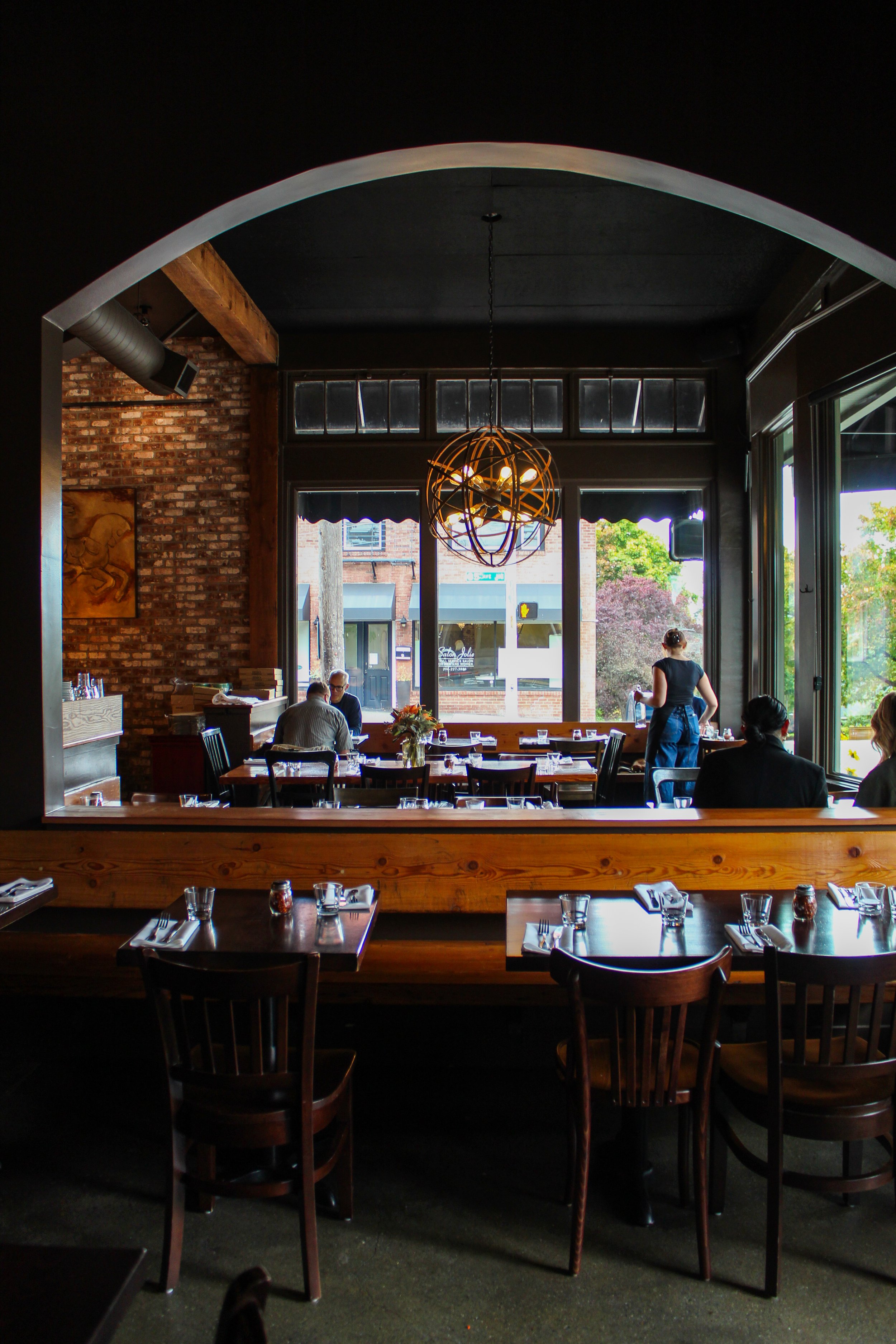 Interior of a restaurant with brick walls and large windows, wooden furniture, and a decorative spherical chandelier hanging from the ceiling. People are seated at tables, and a server is standing nearby.