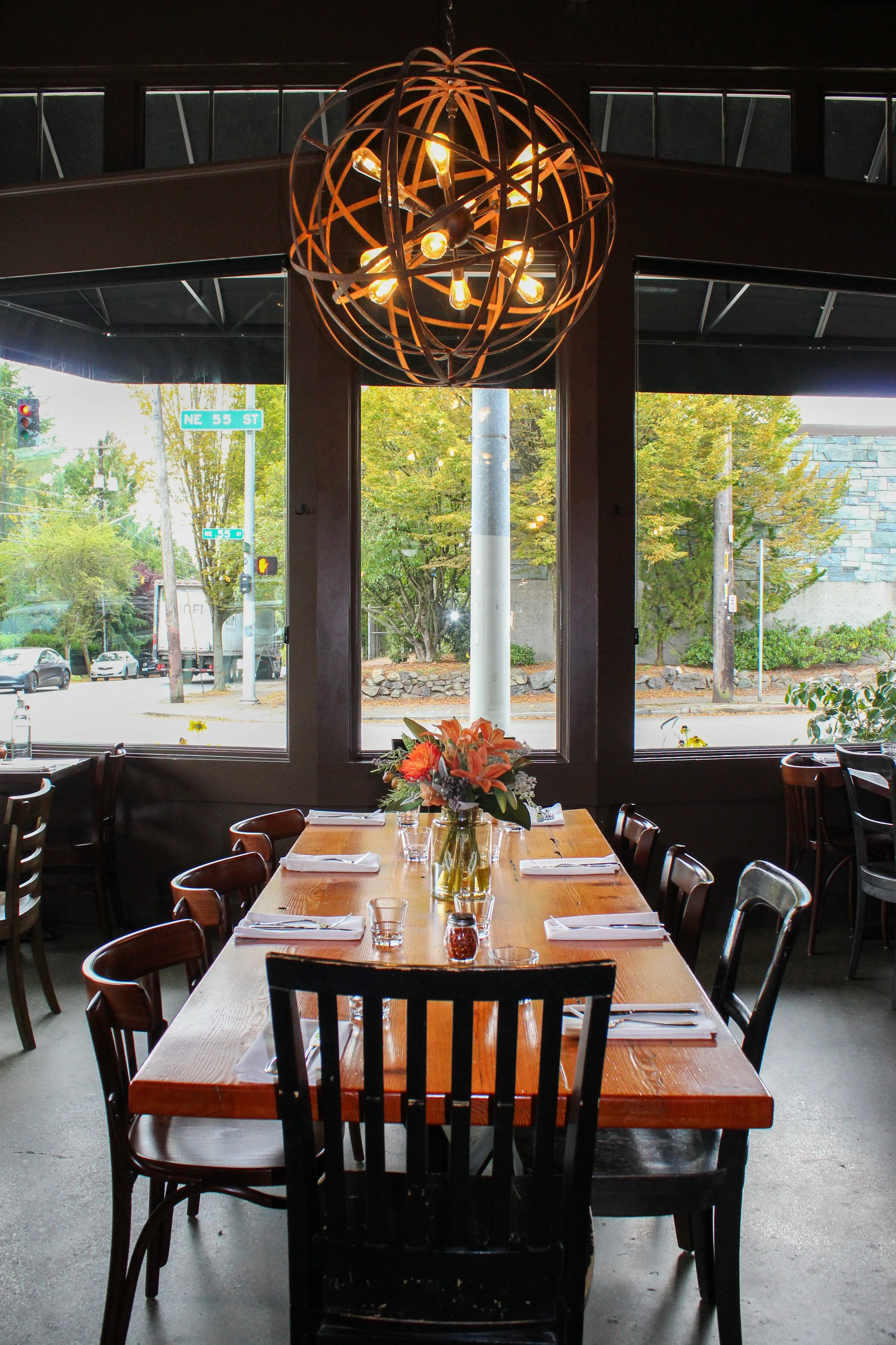 Restaurant interior with a wooden table set with napkins, glasses, and a flower arrangement, and a large globe-shaped chandelier hanging from the ceiling. The window shows a street scene with trees, traffic lights, and street signs reading NE 55 St.
