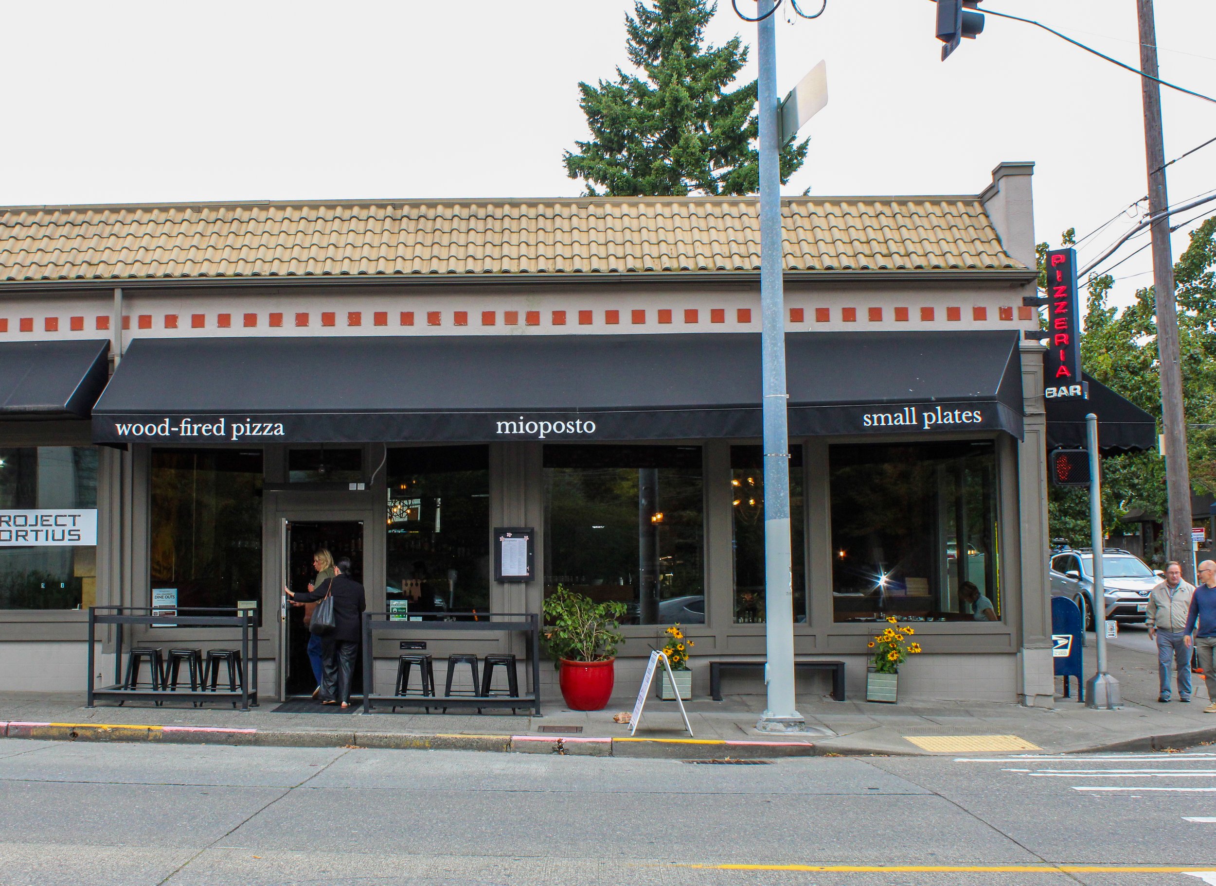 Front view of a restaurant with a beige tiled roof, black awnings, and a sign that reads 'wood-fired pizza, mioposto, small plates'. There are a few people outside, some engaged with their phones, and a large red pot with yellow flowers on the sidewa