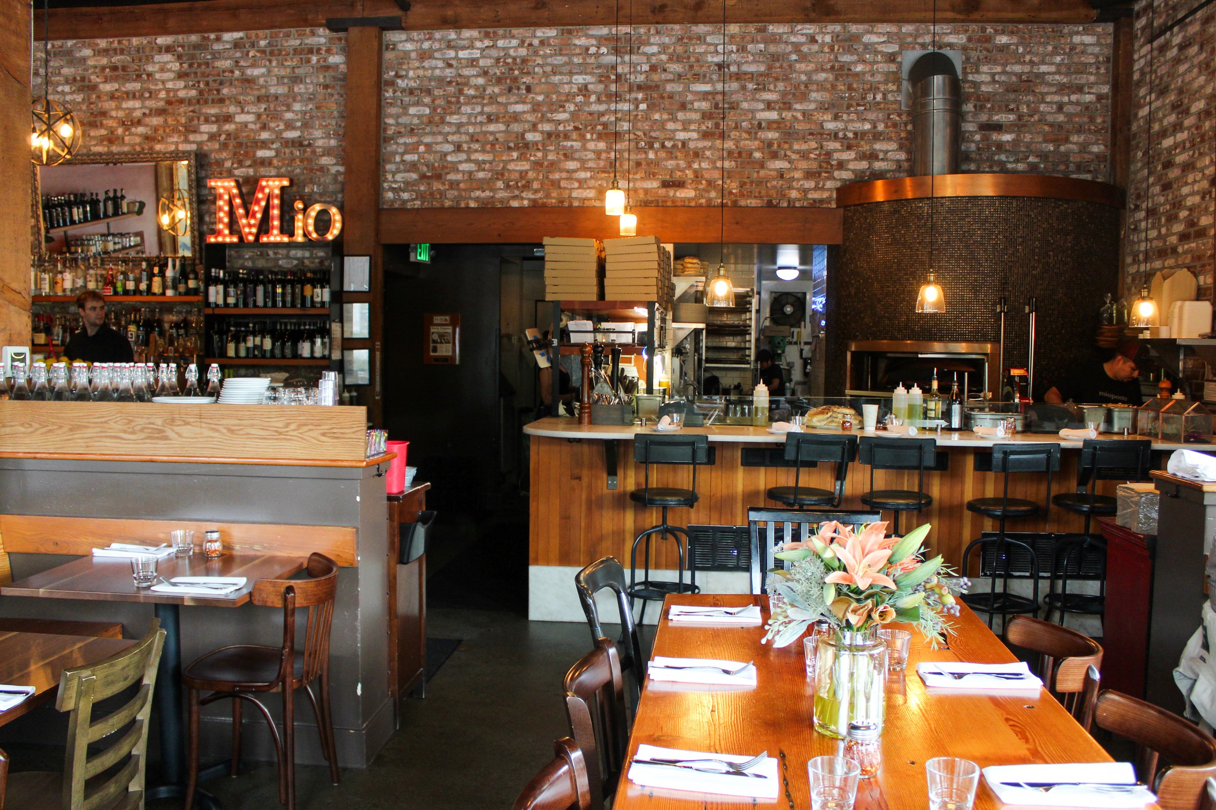 Interior of a restaurant with brick walls, wooden beams, and hanging pendant lights. There is a bar area with bottles and glasses, and a chef working in an open kitchen. The foreground shows a table with a flower arrangement and place settings.