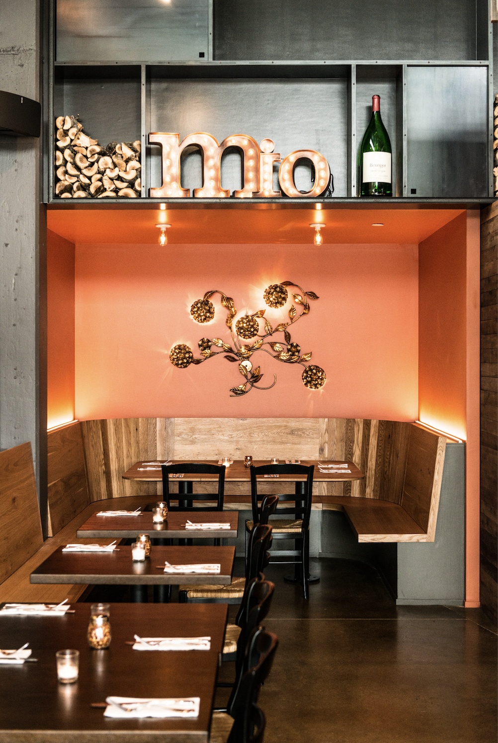 Interior of a restaurant with wooden tables set for dining, a pink wall with a decorative metal art piece, and themed wall decor including a marquee of the word "mio" and a large wine bottle.