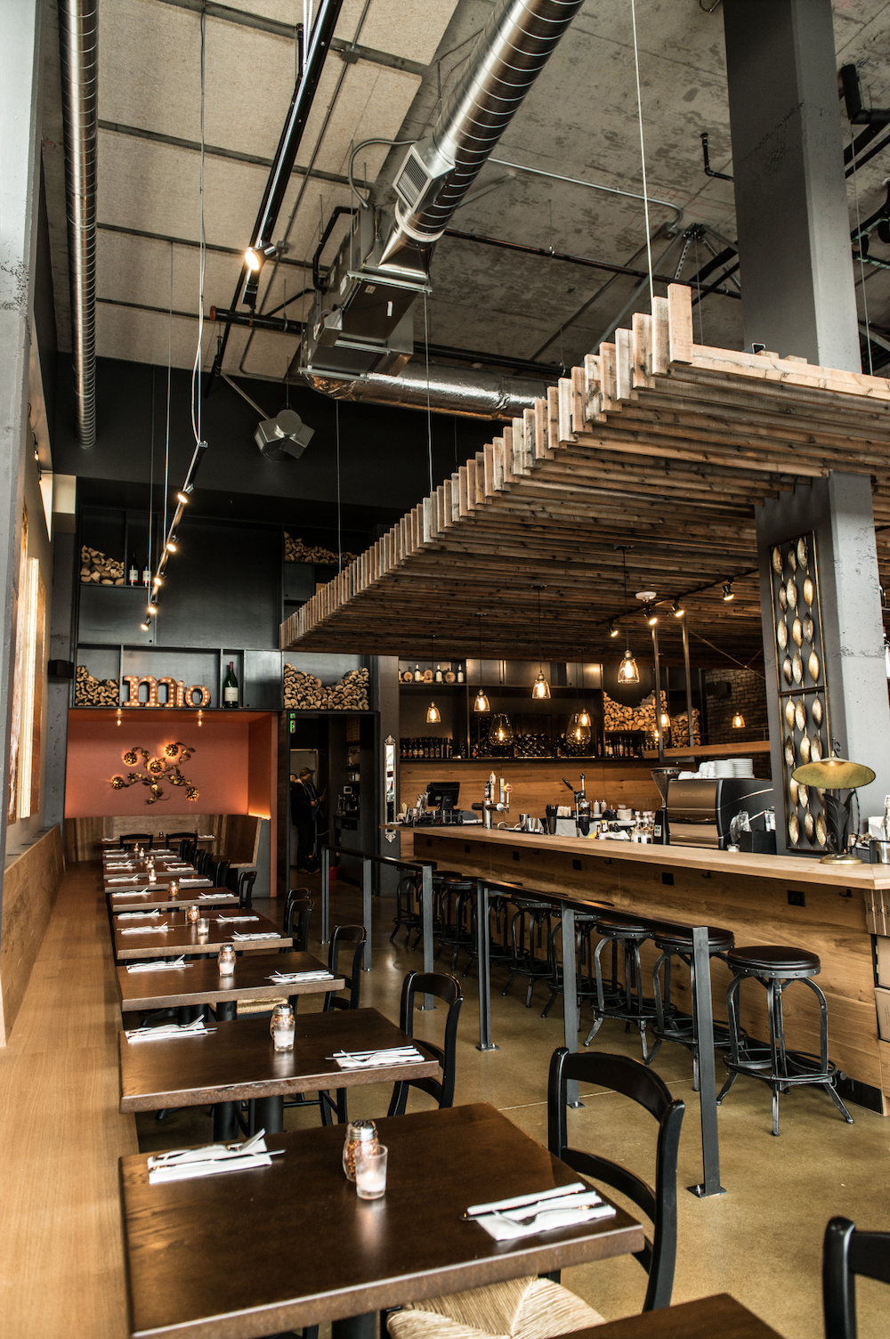 Interior of a modern restaurant with wooden tables and chairs, a bar with stools, black shelving with liquor bottles, and warm ambient lighting.