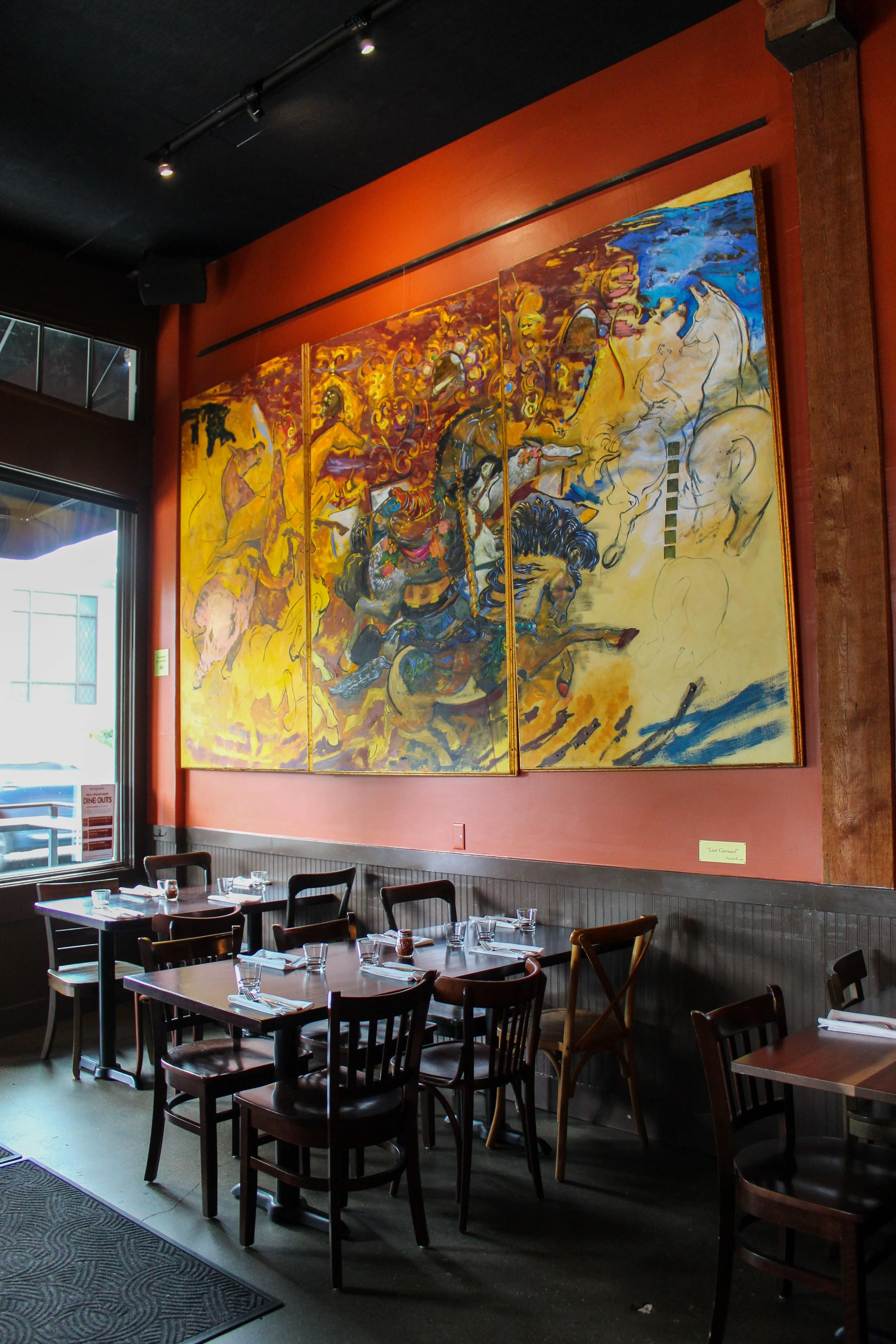 Interior of a restaurant with a large, colorful painting on a reddish wall, tables set with glasses, and chairs arranged for dining.
