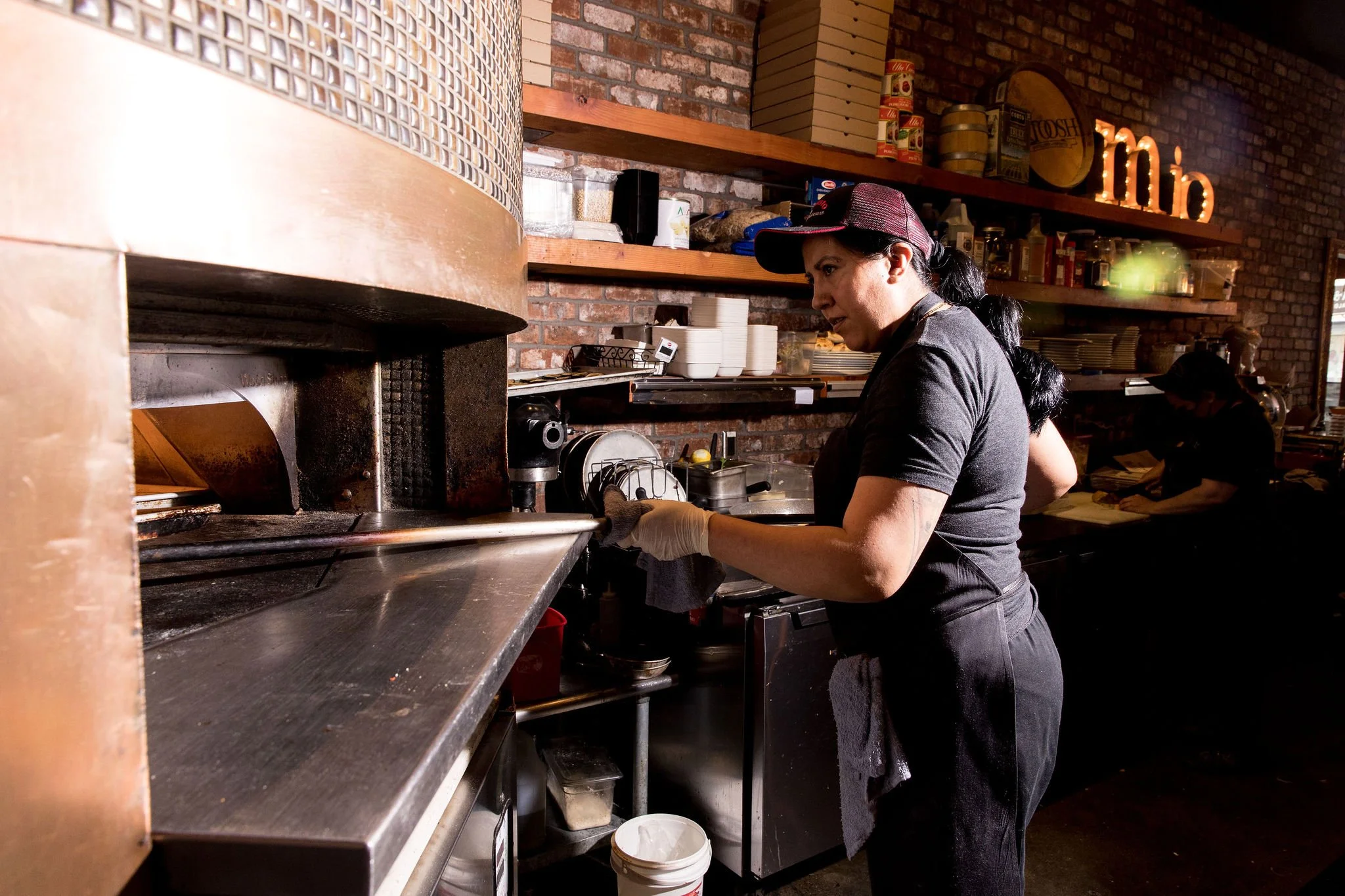 A woman working in a restaurant kitchen, wearing a black shirt, gray apron, and red cap, as she prepares food near a stainless steel counter with shelves and brick walls background.