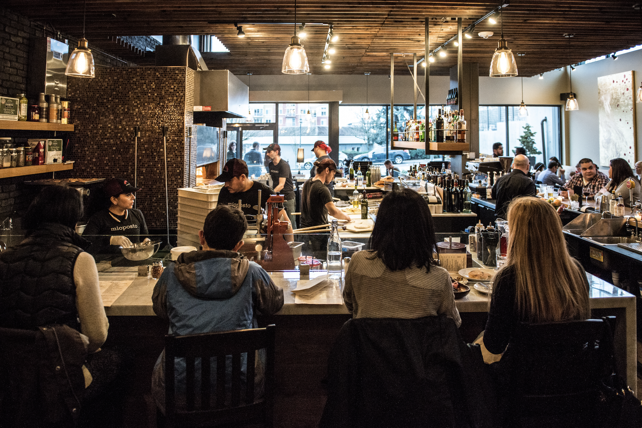 Inside a busy restaurant with customers seated at a bar and tables, staff preparing food, large windows letting in natural light, and warm wooden decor.