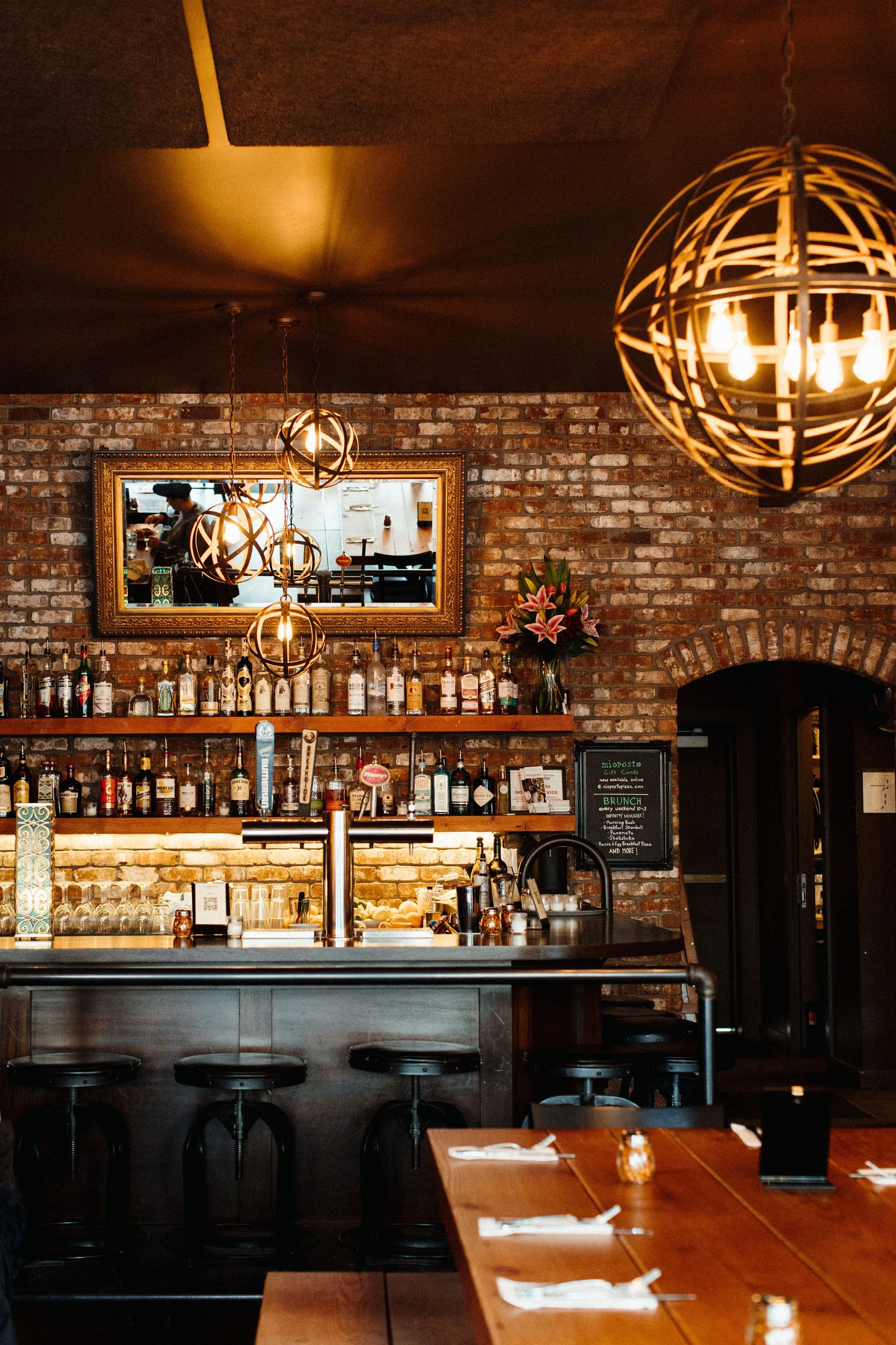 Interior of a bar or restaurant with a brick wall, wooden bar counter, black bar stools, hanging spherical light fixtures, a mirror reflecting a person, and a chalkboard menu.