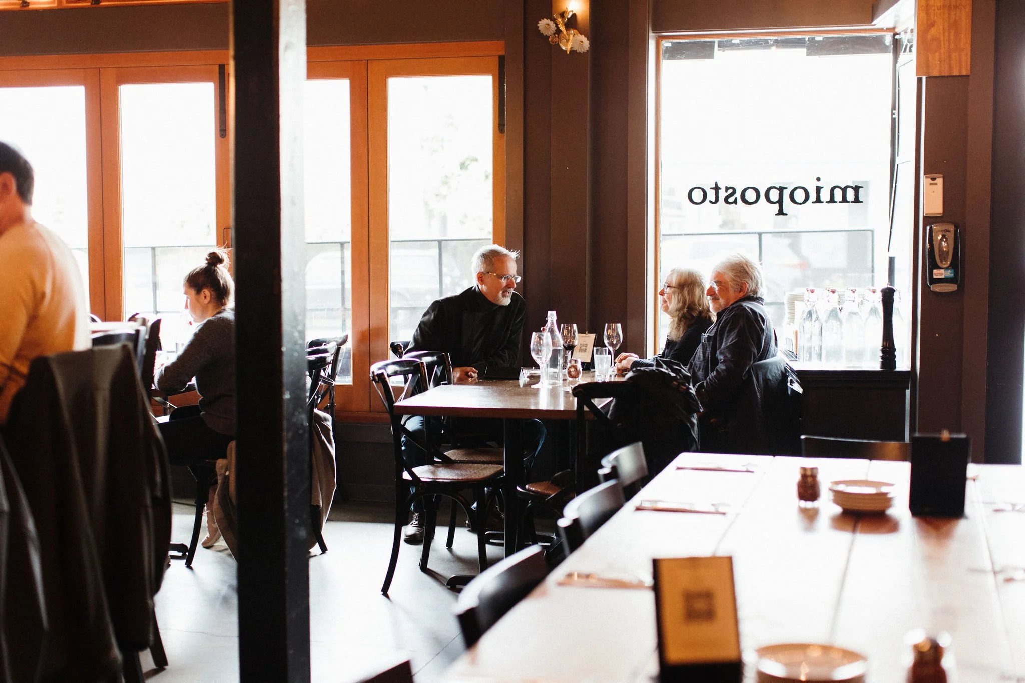 People dining inside a restaurant with large windows, tables, and chairs. The window has a reflection of the restaurant's name, 'mipoosta'.