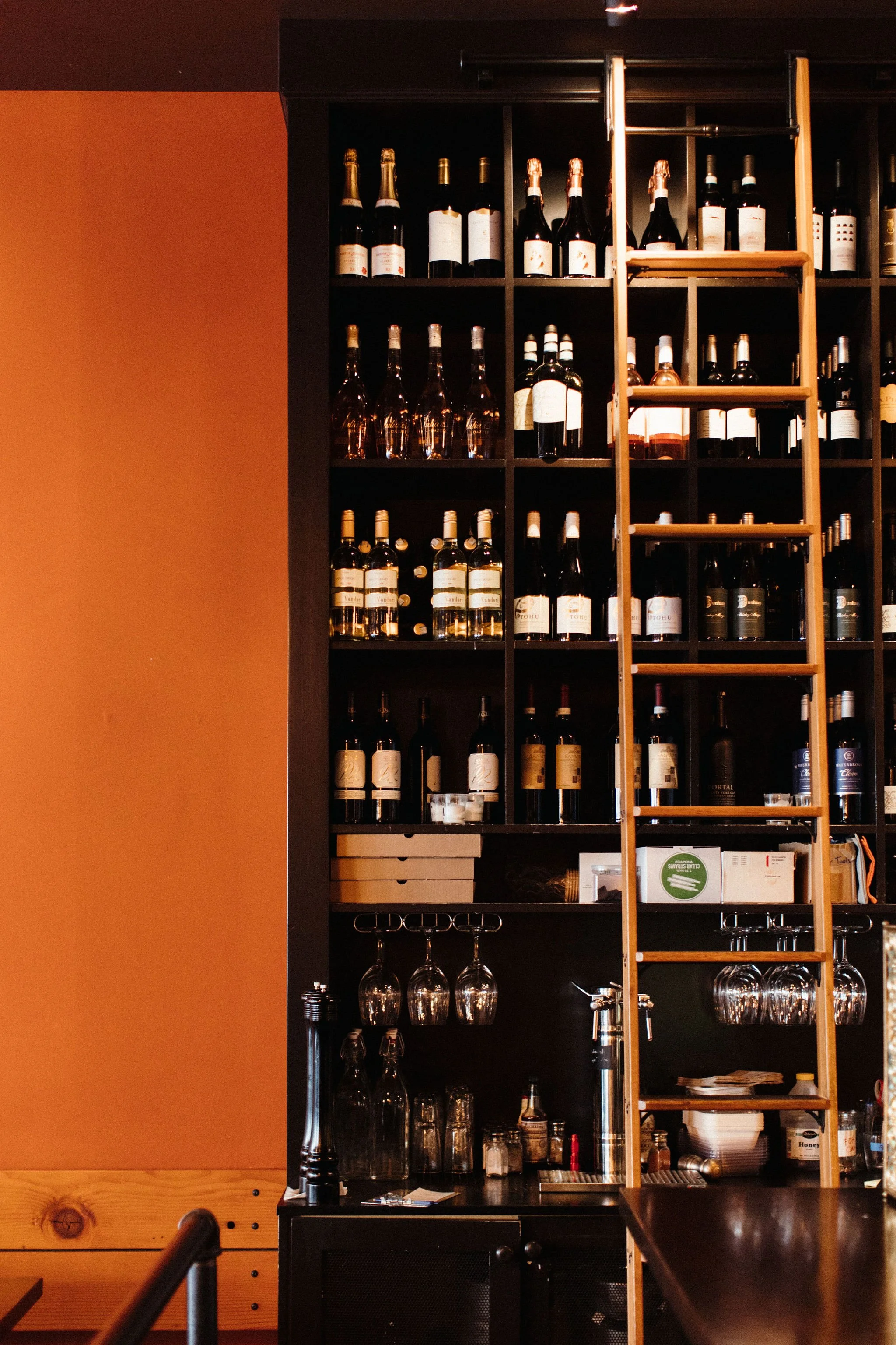 Shelf filled with various bottles of alcohol, wine, and liquor behind a bar. There is a wooden ladder leaning on the shelf, and some glassware is hanging below.