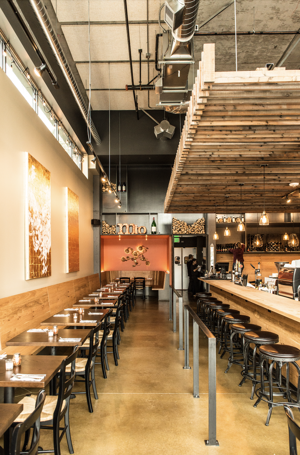 Interior of a modern restaurant with a bar counter on the right, wooden tables with chairs on the left, warm lighting, artwork on the walls, and a rustic wood ceiling.