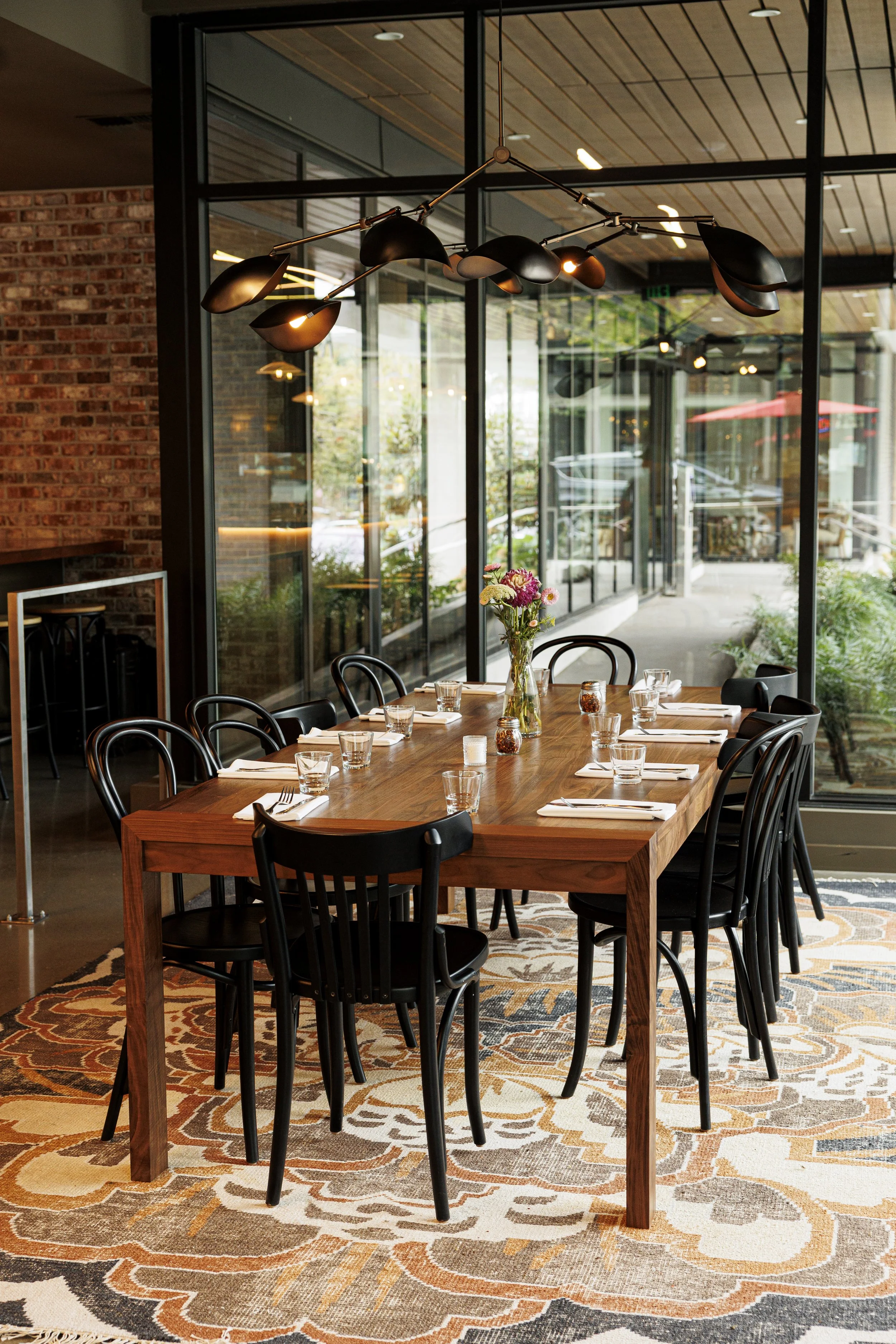 A dining table set with glassware, napkins, and a flower arrangement in a vase, surrounded by black chairs. The room features large glass windows, a patterned rug, and a modern pendant light fixture overhead.