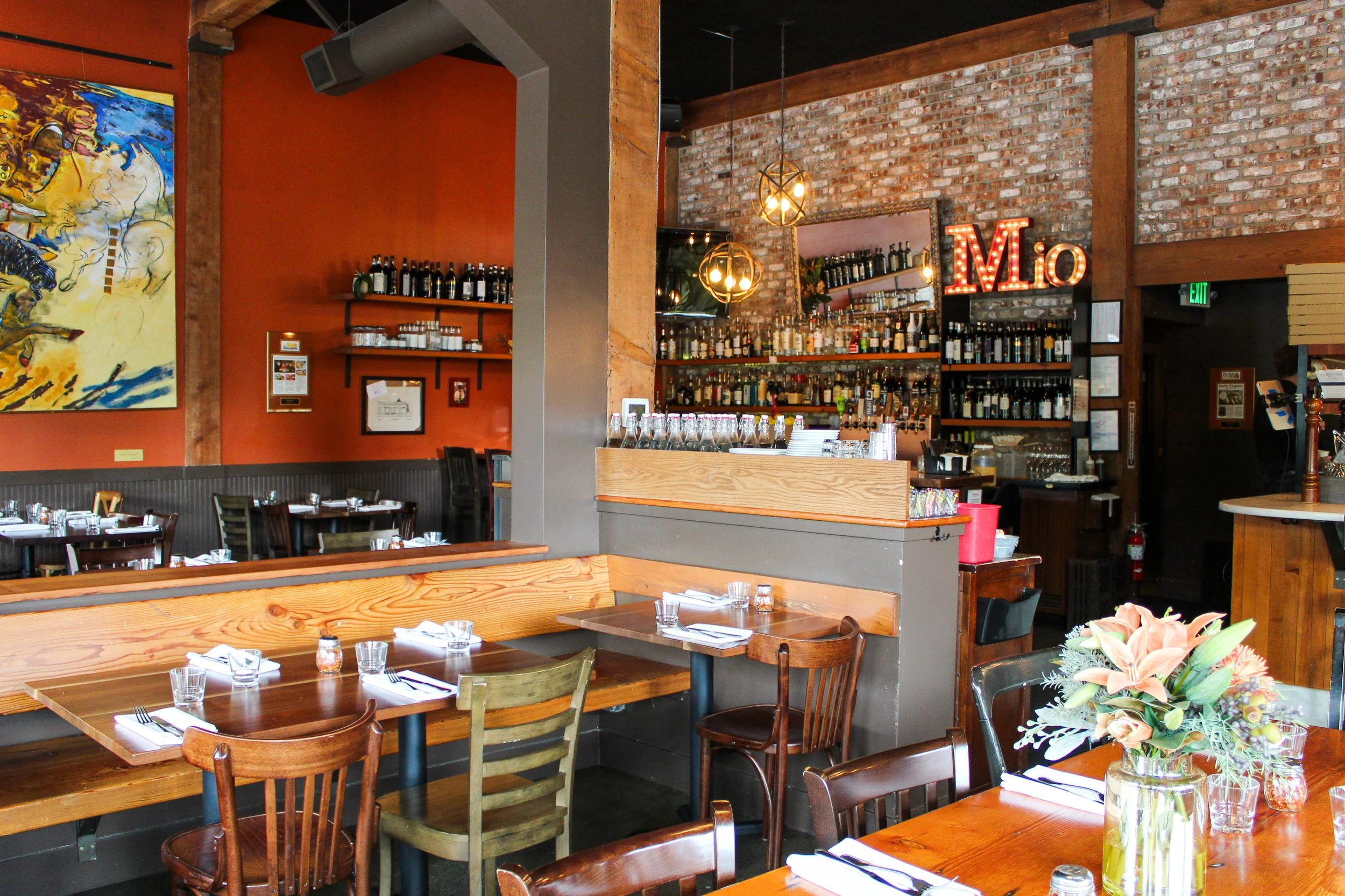 The interior of a restaurant with wooden tables and chairs, exposed brick walls, a bar area with bottles, and decorative lighting fixtures.