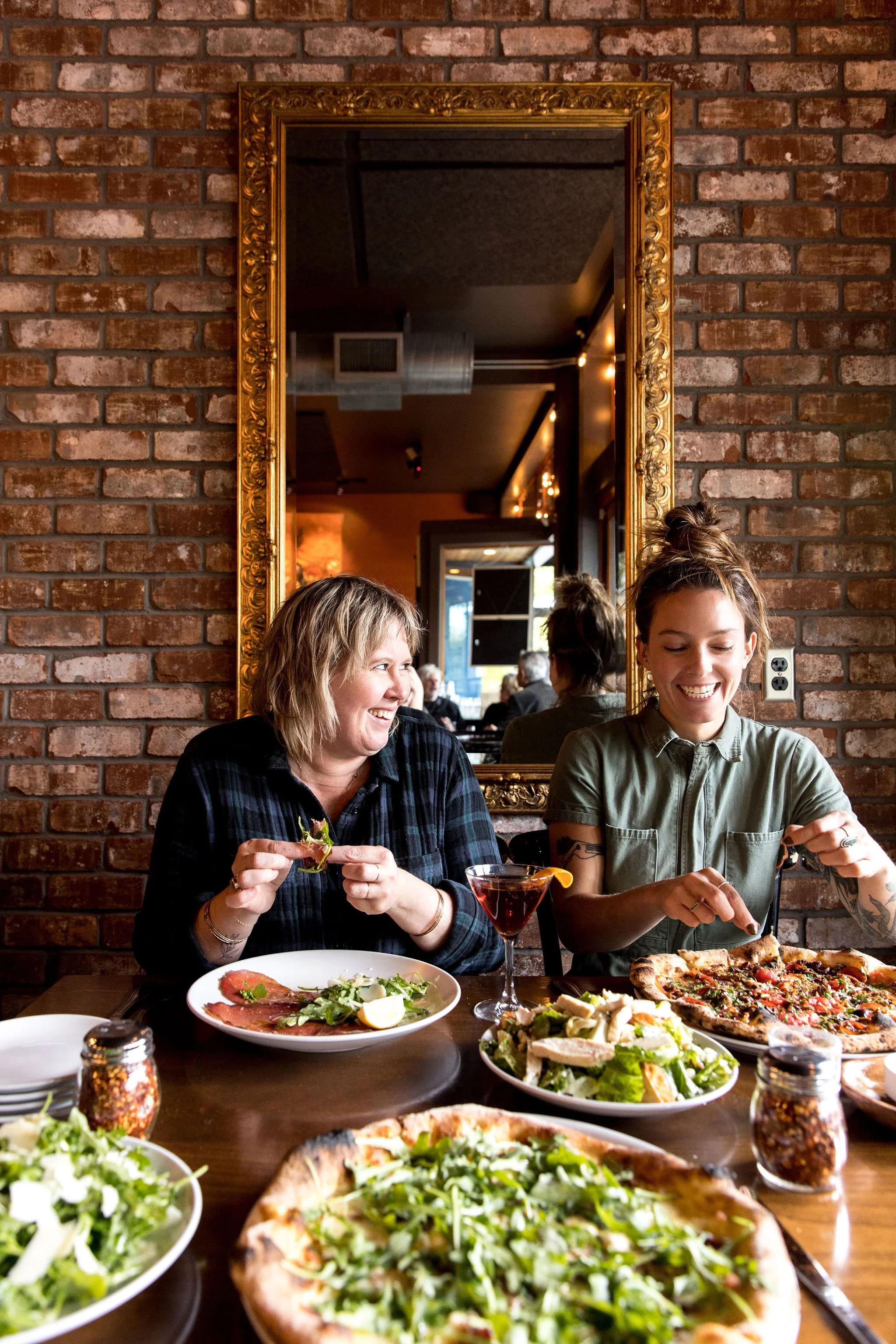 Two women sitting at a table with pizza, salad, and drinks in a restaurant with brick walls and a large mirror.
